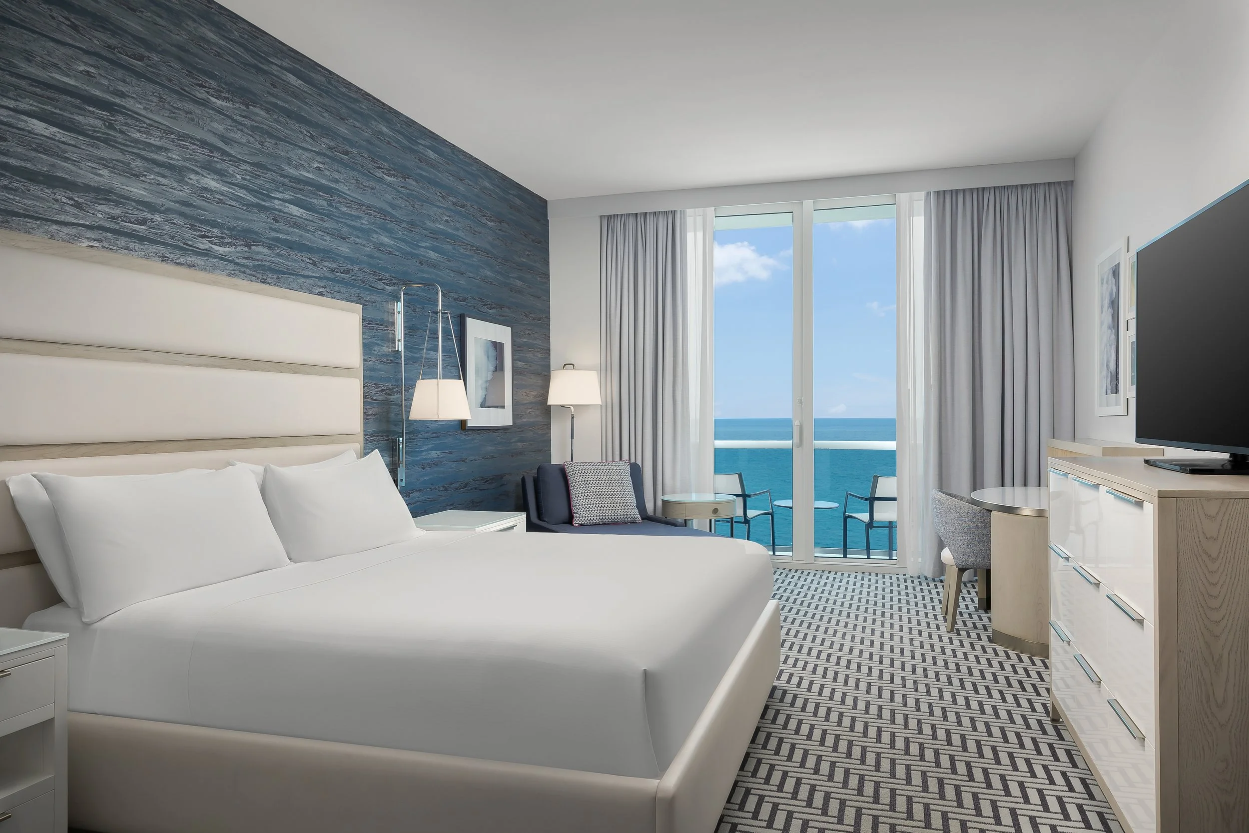 Modern hotel room with white bed, blue accent wall, large window showing ocean view, sitting area with chairs, and television.