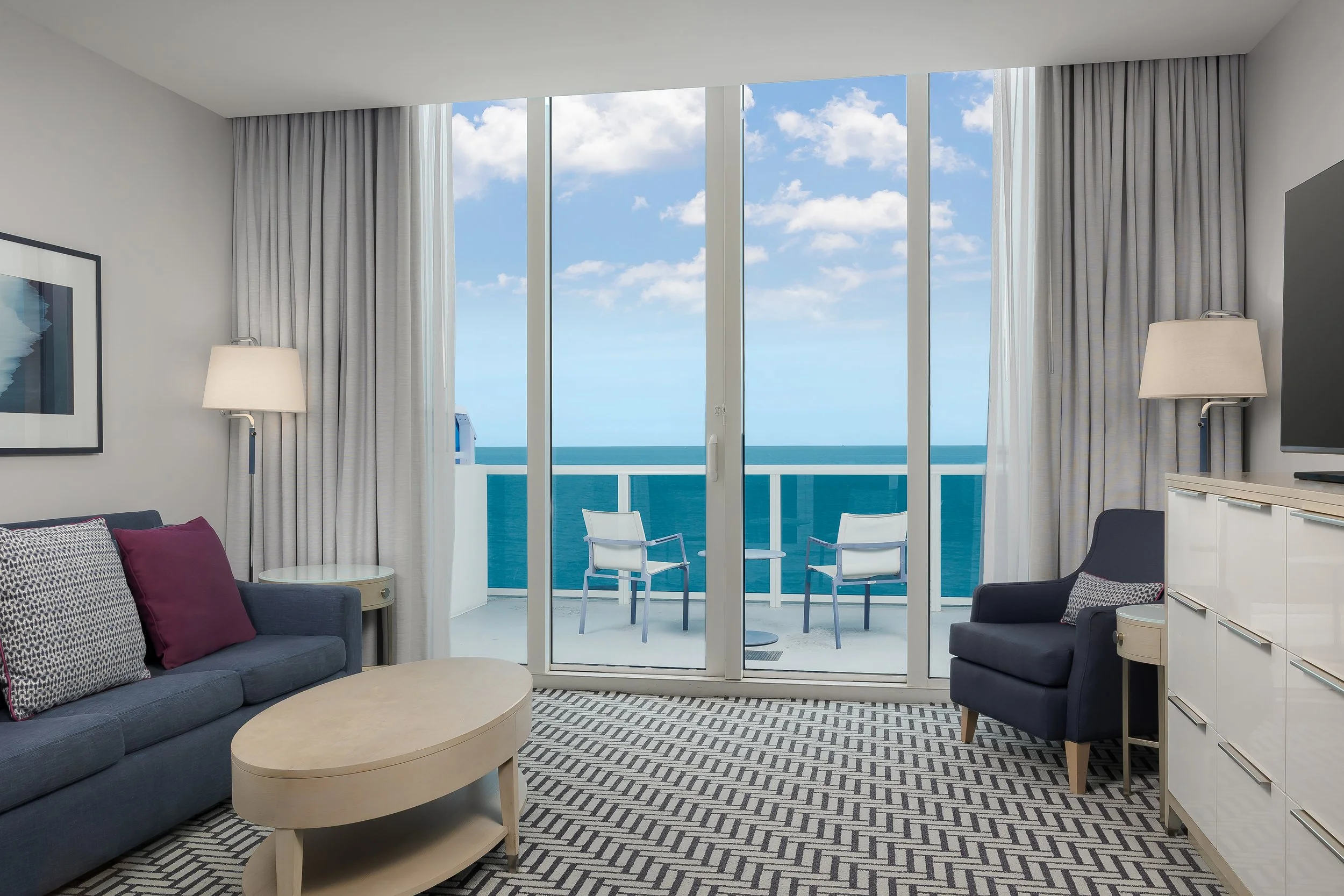 Living room with large windows overlooking the ocean, furnished with a gray sofa with purple pillows, a beige oval coffee table, and a black armchair. There are two white lamps and a framed artwork on the wall.