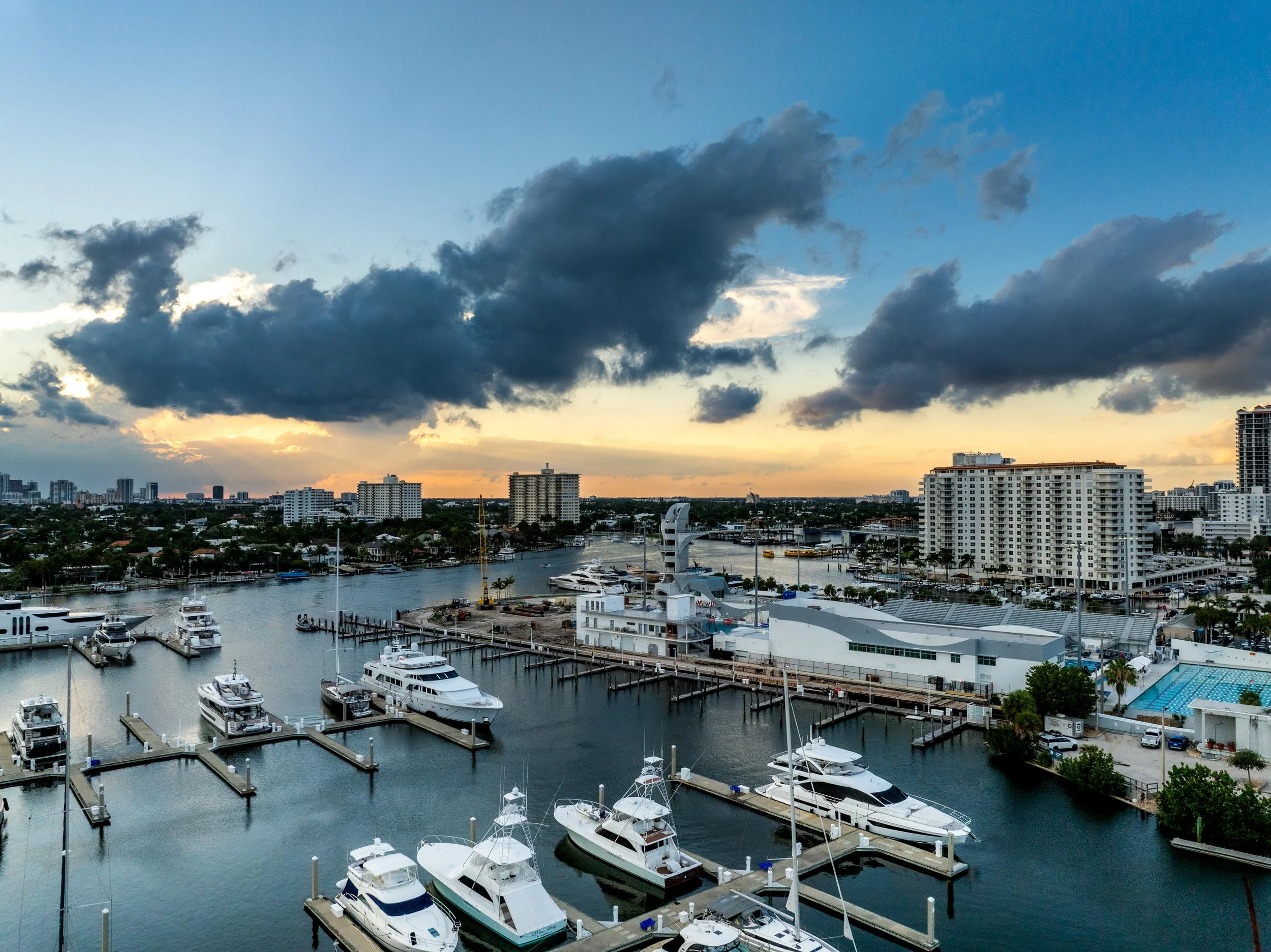 A cityscape view of a marina with boats docked, high-rise buildings in the background, and a sky with dark clouds at sunset.