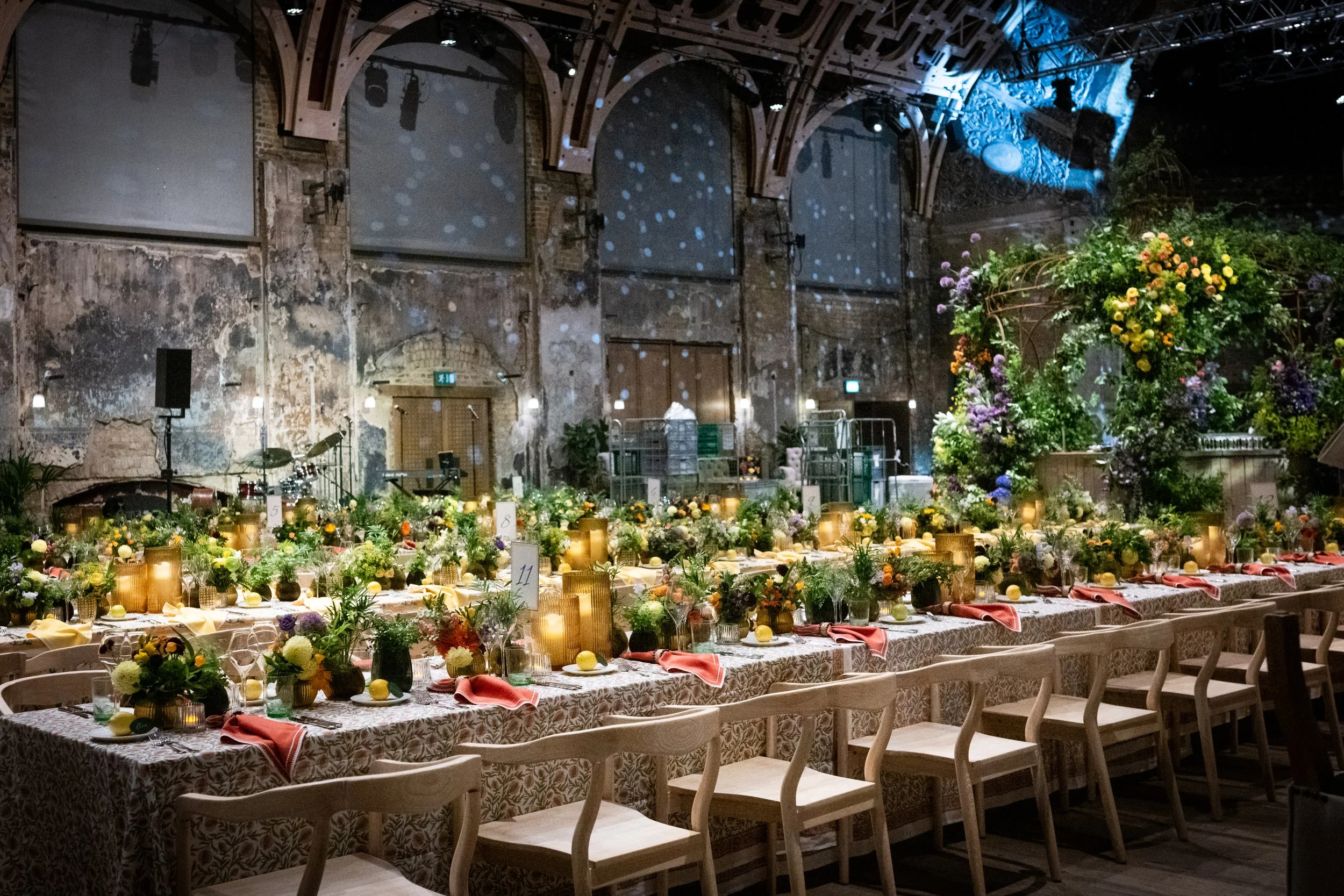 An indoor event space with rustic brick and wooden beams decorated with string lights, set up for a wedding or celebration. Long tables are adorned with floral arrangements, candles, and tableware, and a large, colorful floral centerpiece is on one e