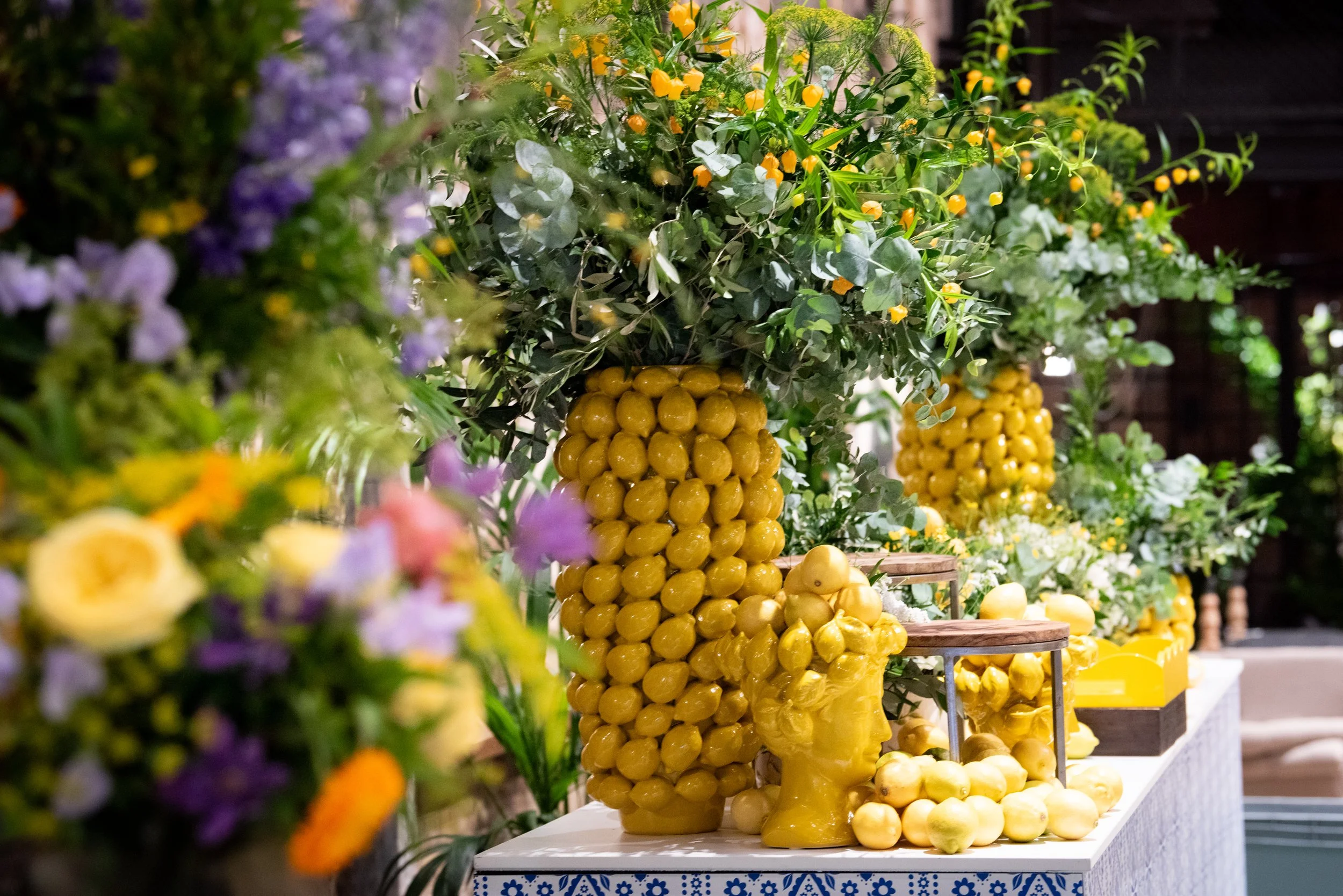 Display of lemons on a table with decorative yellow bases, surrounded by green foliage and purple flowers in an indoor setting.