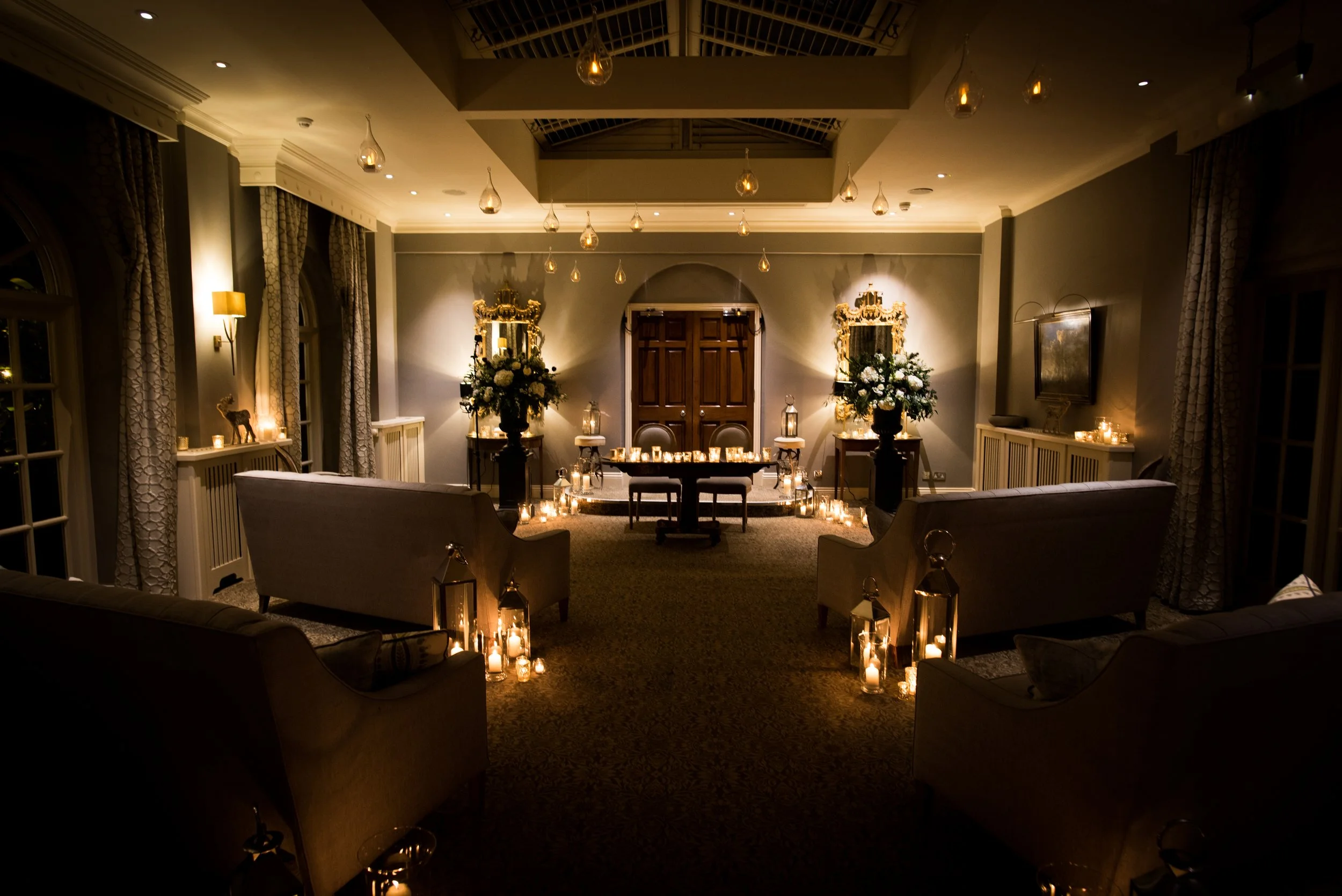 A beautifully decorated, dimly lit room with elegant sofas, floral arrangements, and numerous candles creating a romantic ambiance, likely prepared for a special event or celebration.
