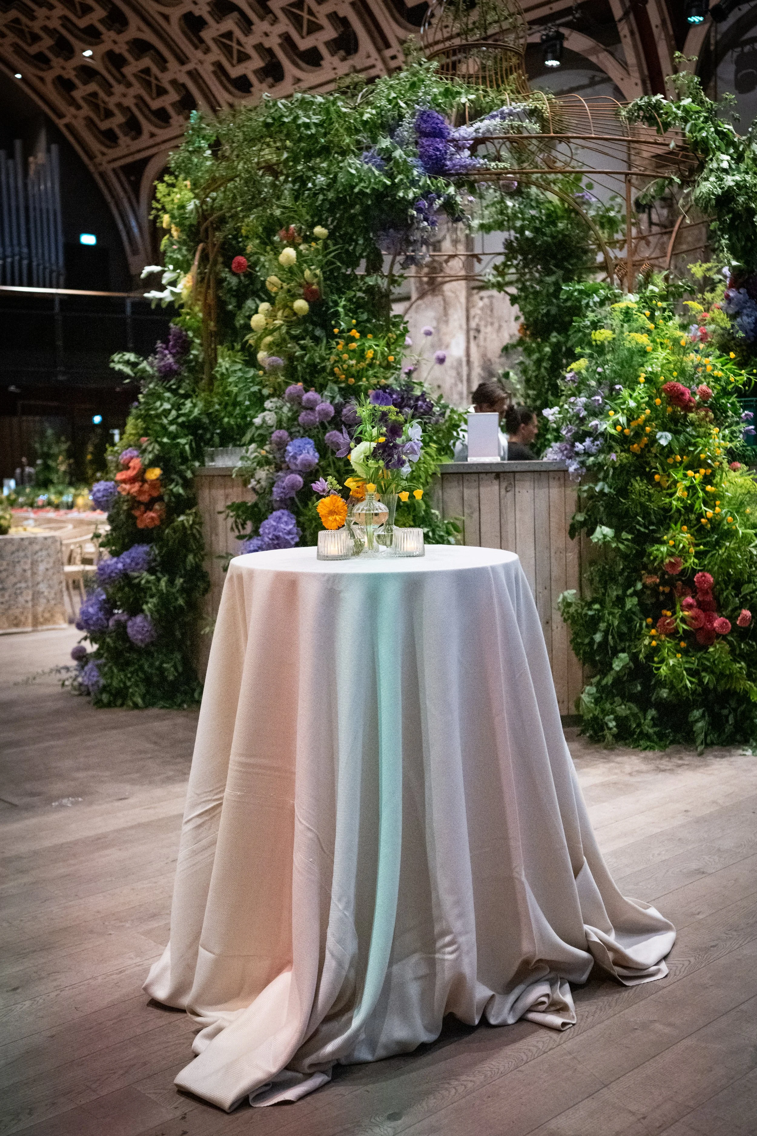 A round table covered with a white tablecloth, set with vases containing purple, yellow, and orange flowers, in an indoor space with wooden floors, surrounded by a floral arch and greenery in the background.