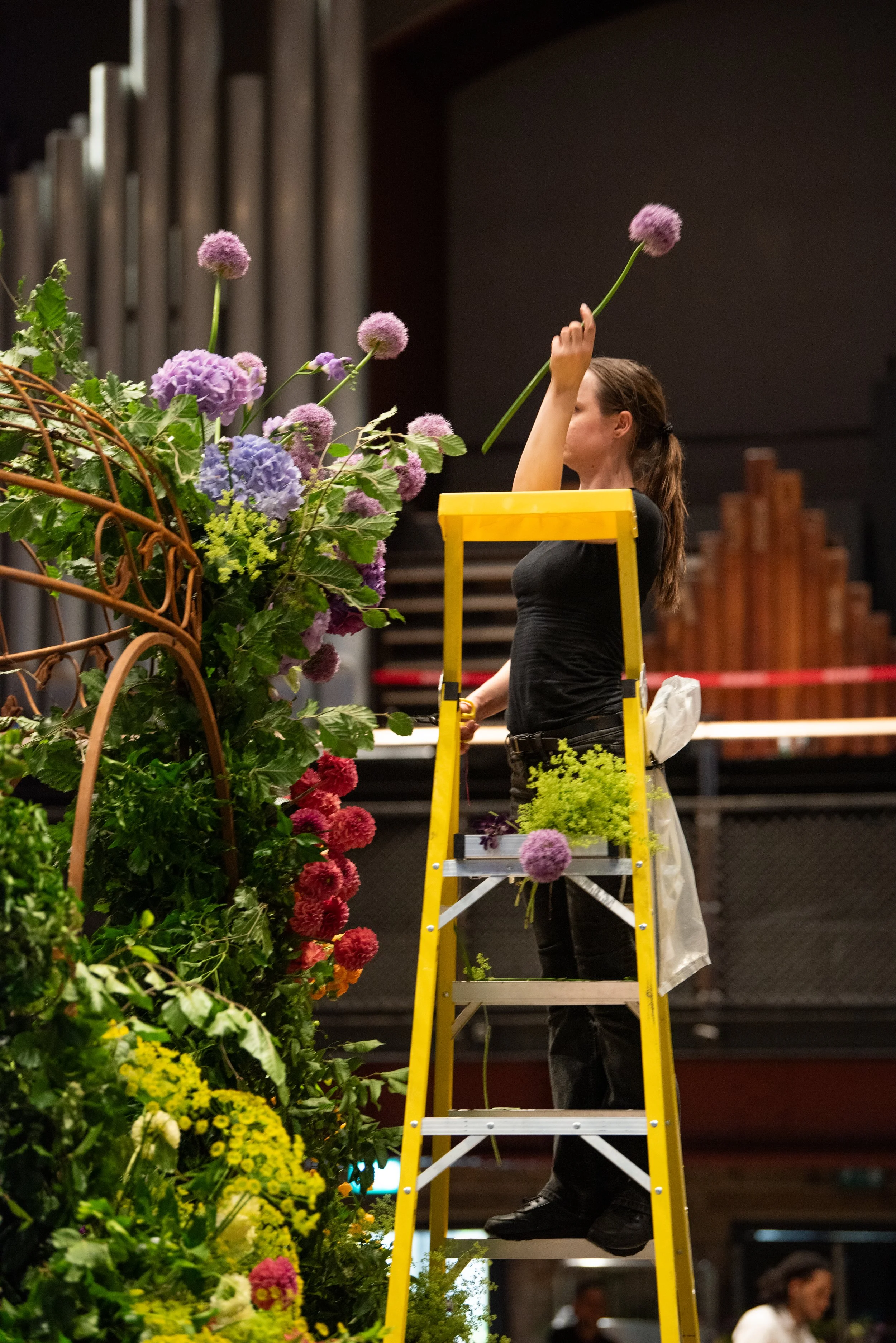 A woman standing on a yellow step ladder arranging purple and pink flowers on a floral display.