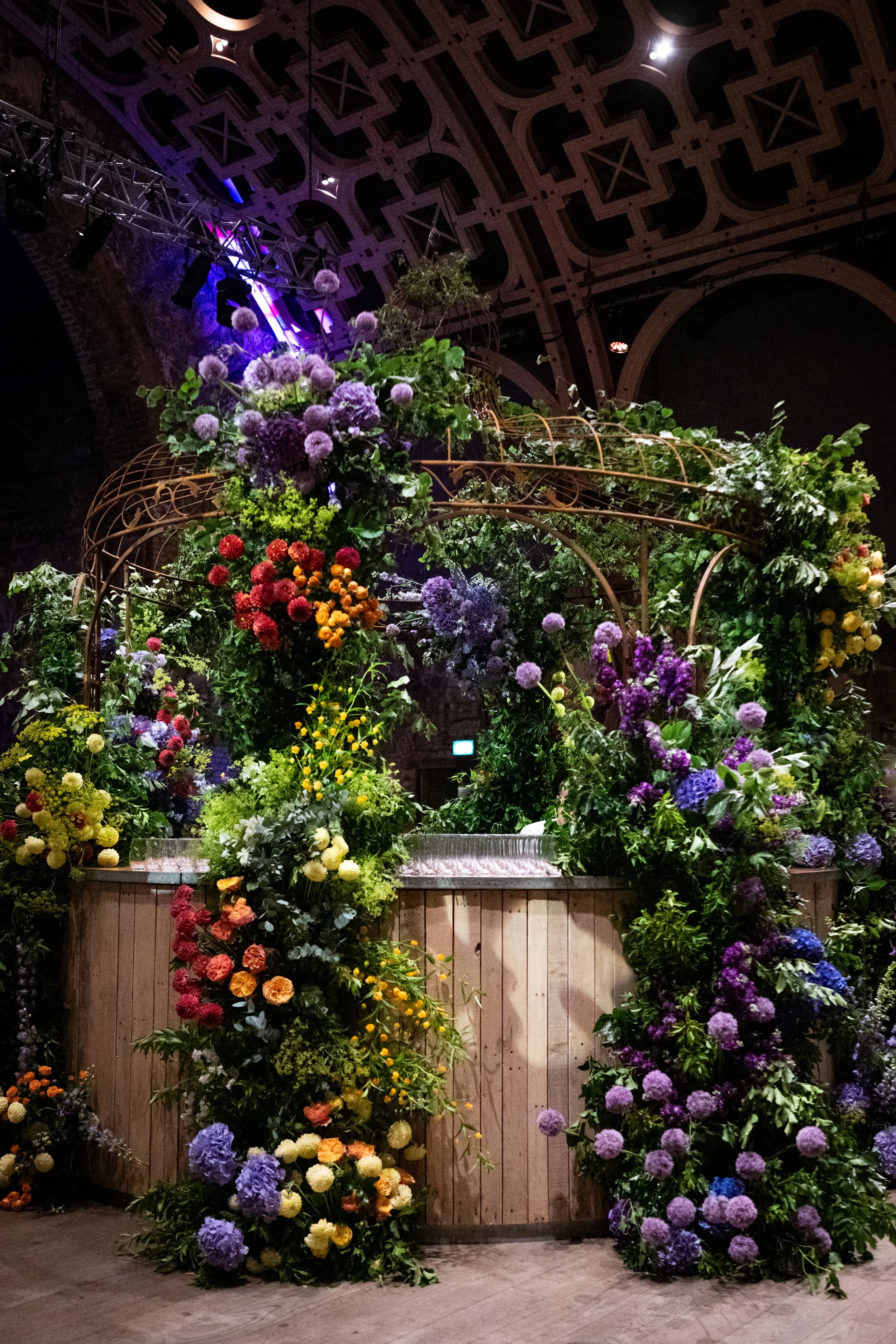 A stage decorated with a variety of colorful flowers arranged on wooden structures and the floor, with a dark ceiling featuring geometric patterns and stage lighting.