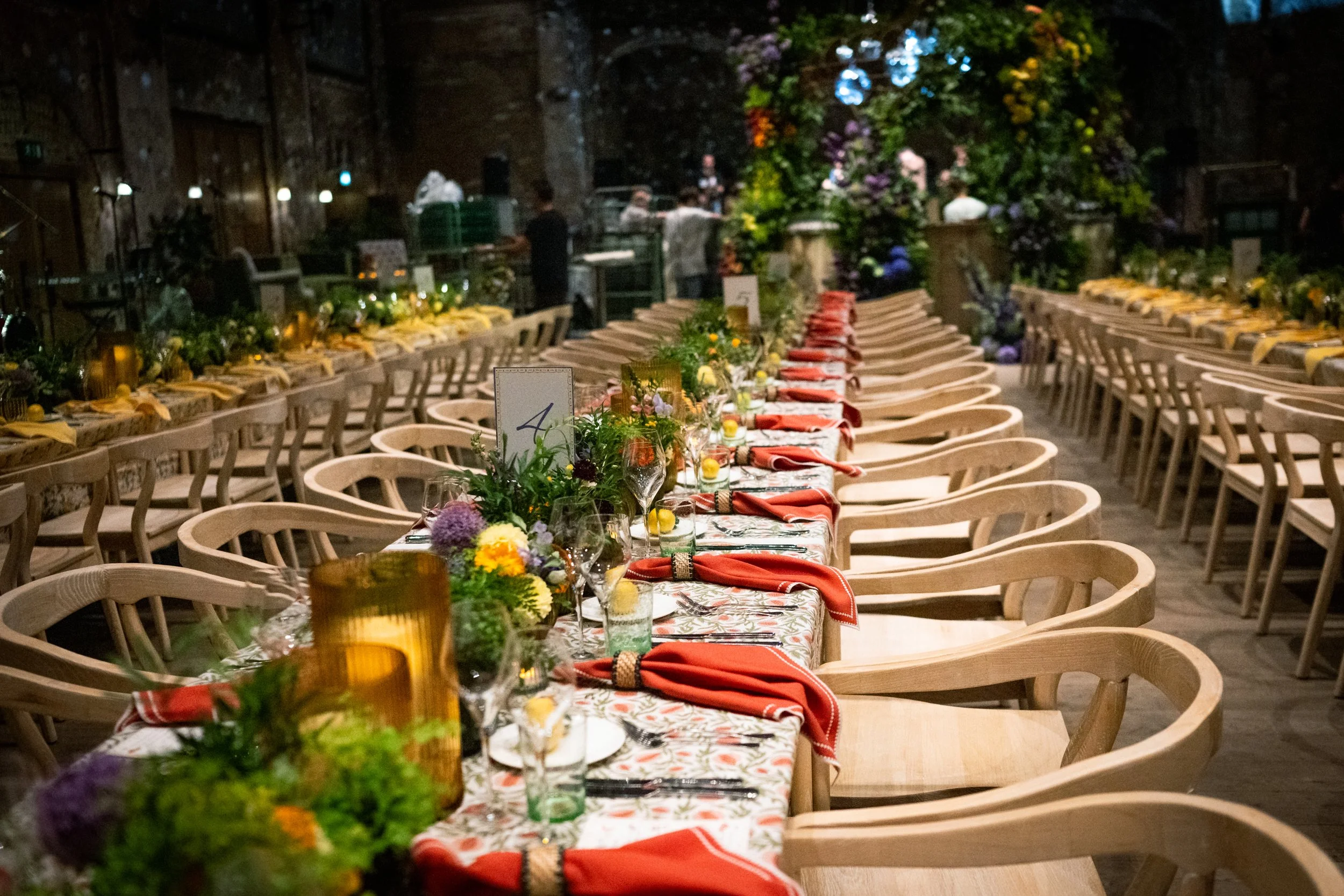 Long dining table set for an event with floral centerpieces, glasses, napkins, and plates in an industrial-style venue decorated with greenery and flowers.