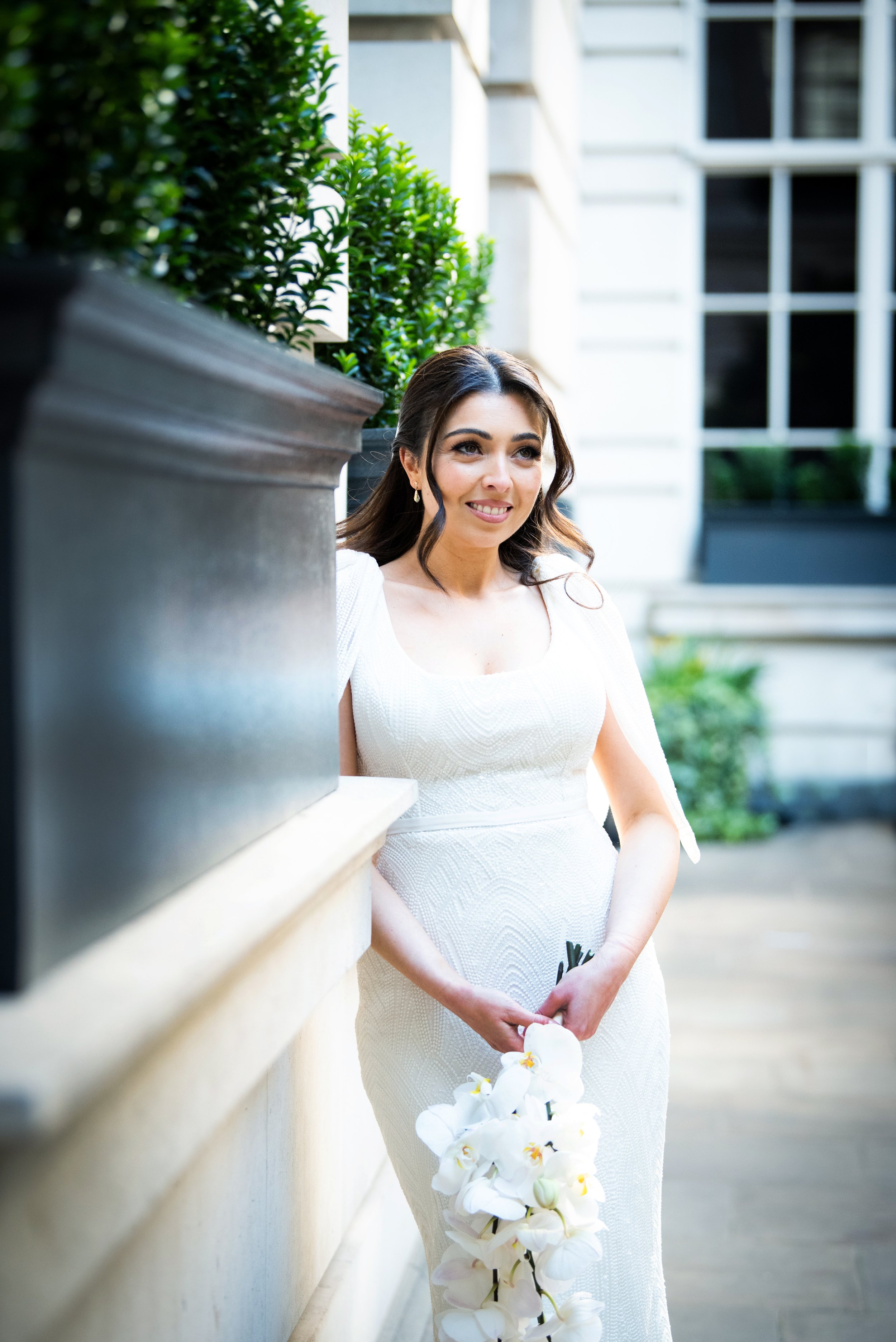 Black and white photo of a couple walking hand in hand on a city street, with a large ornate building on the right and potted plants along the sidewalk.