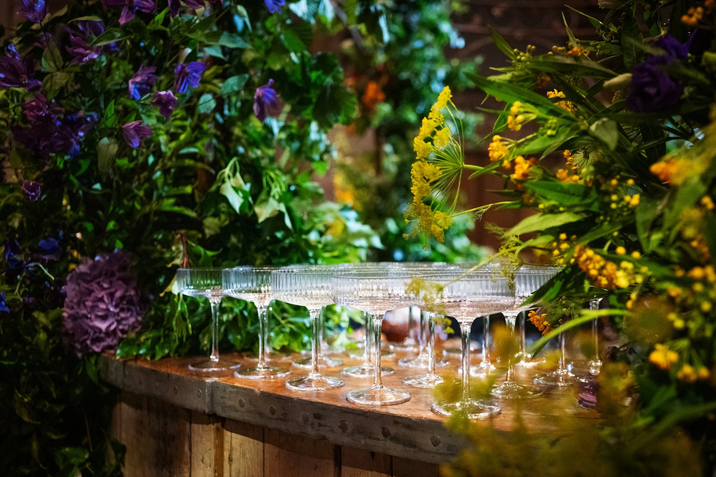 A table with several empty champagne glasses surrounded by a lush arrangement of colorful flowers and greenery, creating an elegant and vibrant setting.