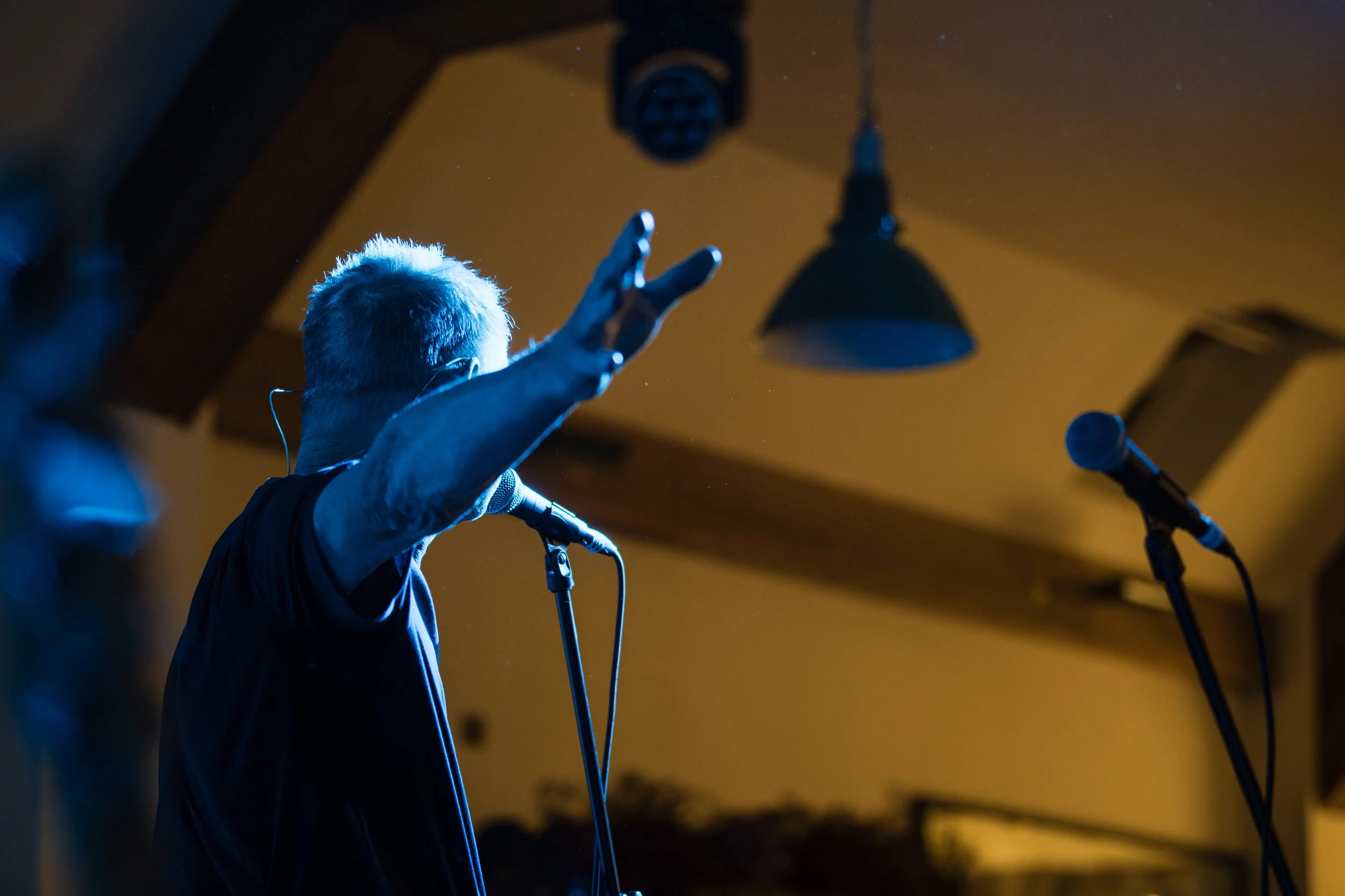 A musician with platinum blonde hair performing on stage with a raised hand, illuminated by blue stage lights, with microphones visible in front.