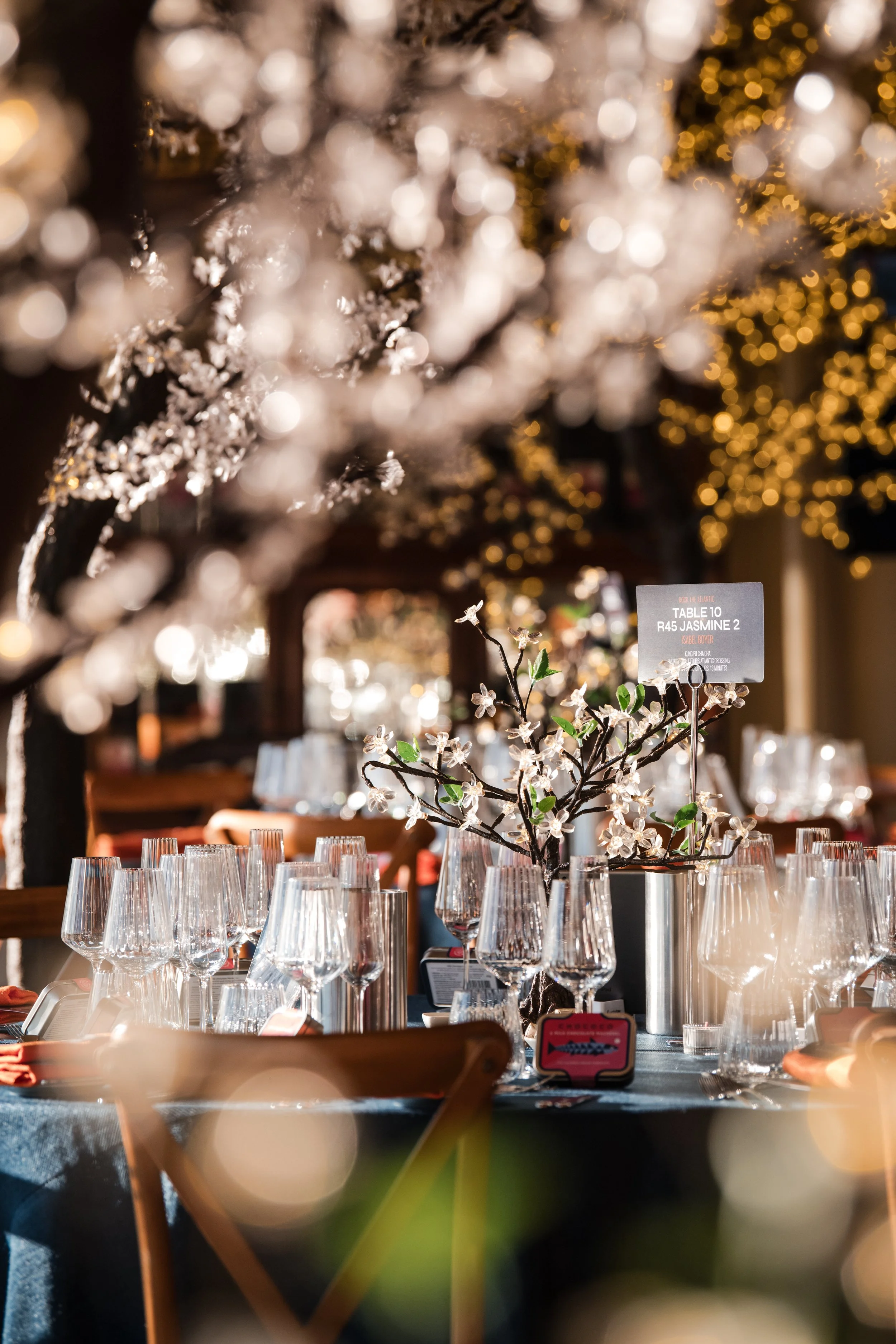 A decorated table set for a formal gathering with numerous empty wine glasses, a centerpiece with white flowers and green leaves, and a small sign indicating seating arrangements. The background features twinkling yellow and white lights, creating a 