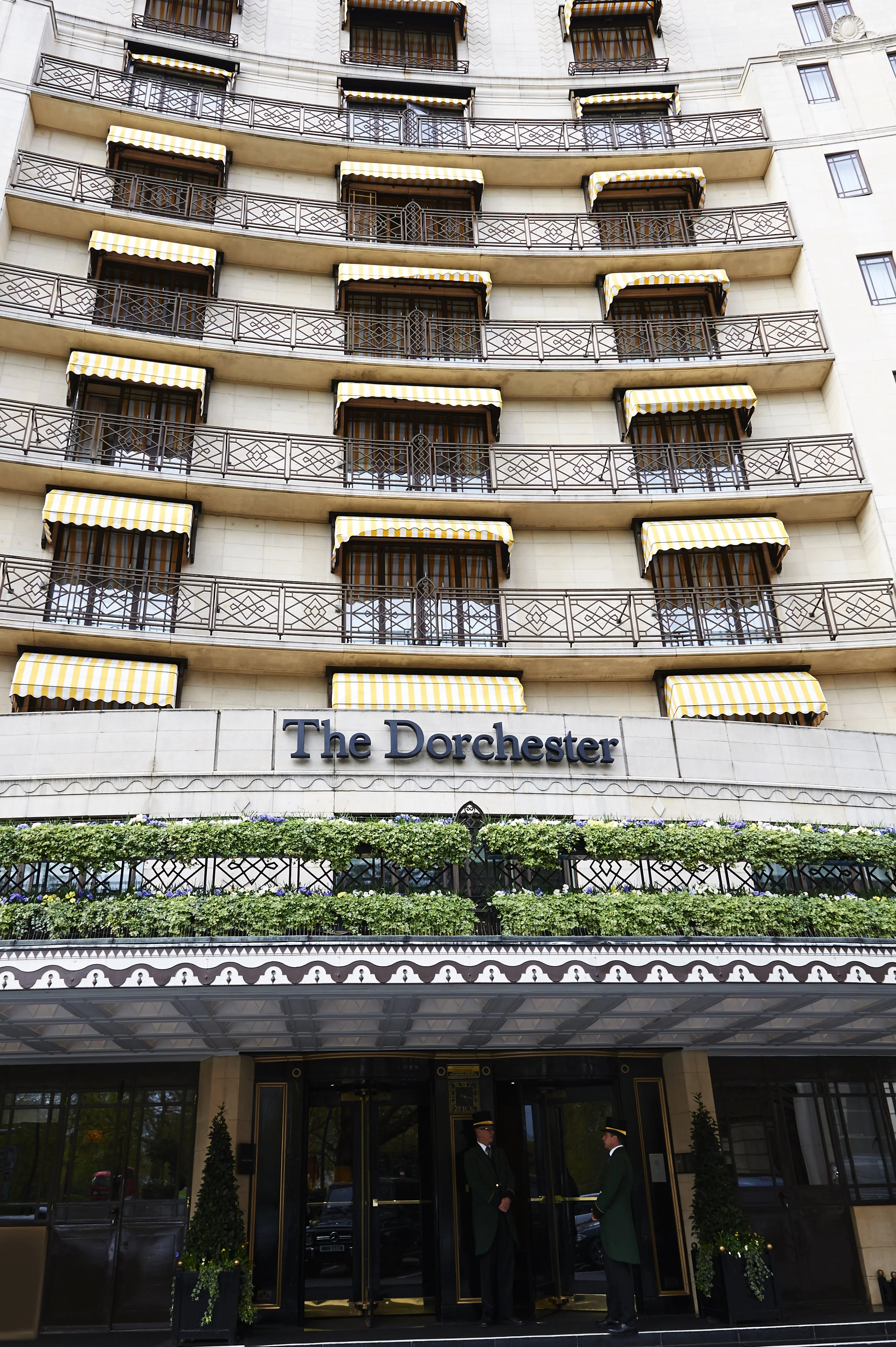 The facade of The Dorchester hotel with multiple balconies with yellow and white striped awnings, and two doormen standing at the entrance.