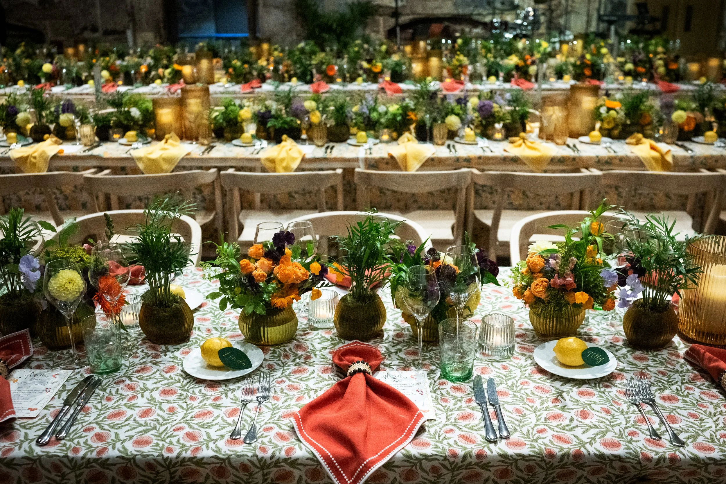 A long banquet table decorated with numerous floral arrangements in vases, set for a formal event with place settings, glasses, napkins, and small yellow lemons, in an indoor venue.