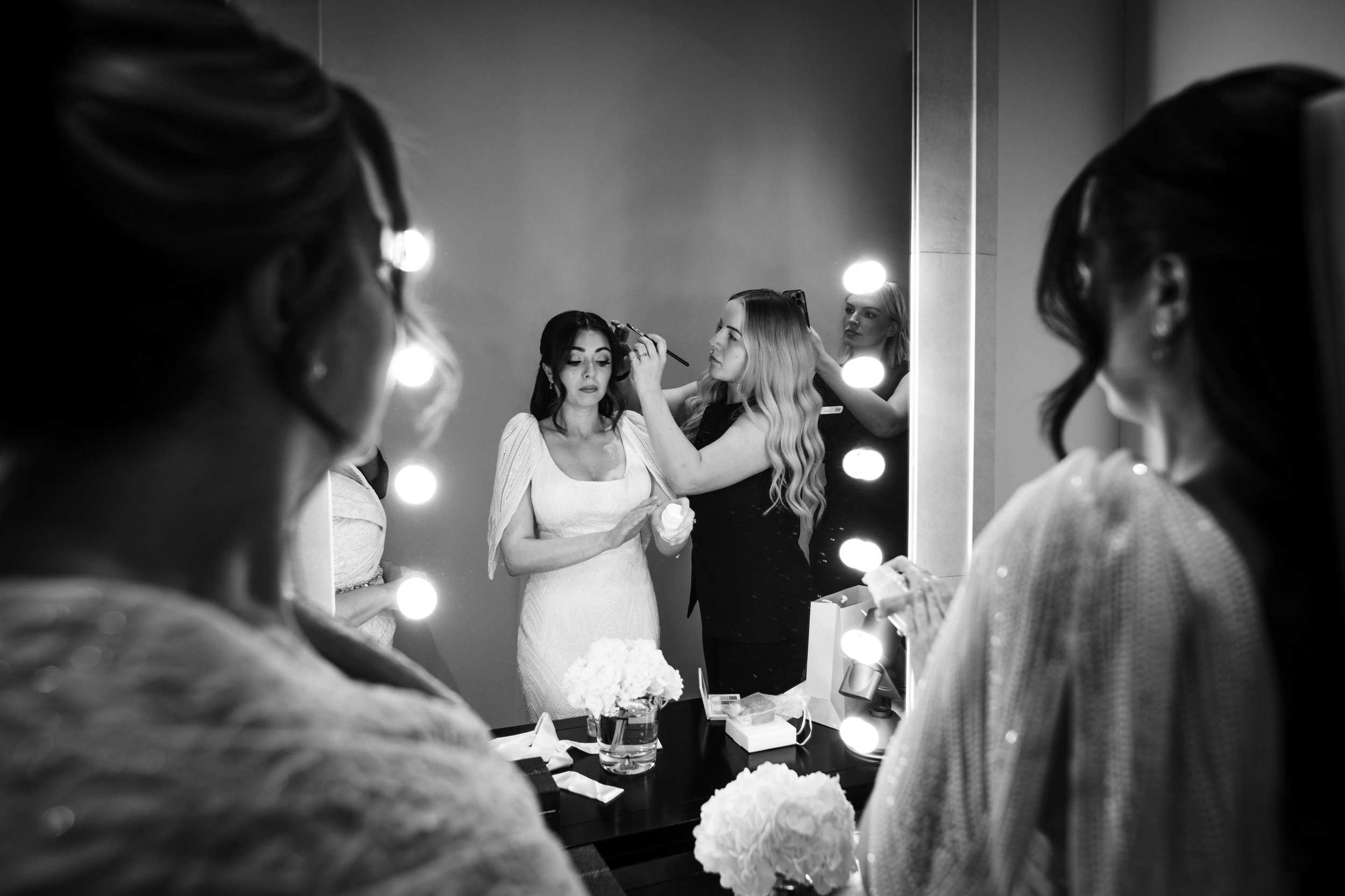A woman in a white dress, likely a bride, is having makeup applied by another woman. They are in front of a mirror with bright round lights, possibly backstage or in a dressing room, and there are other women and a table with a flower arrangement and makeup items.