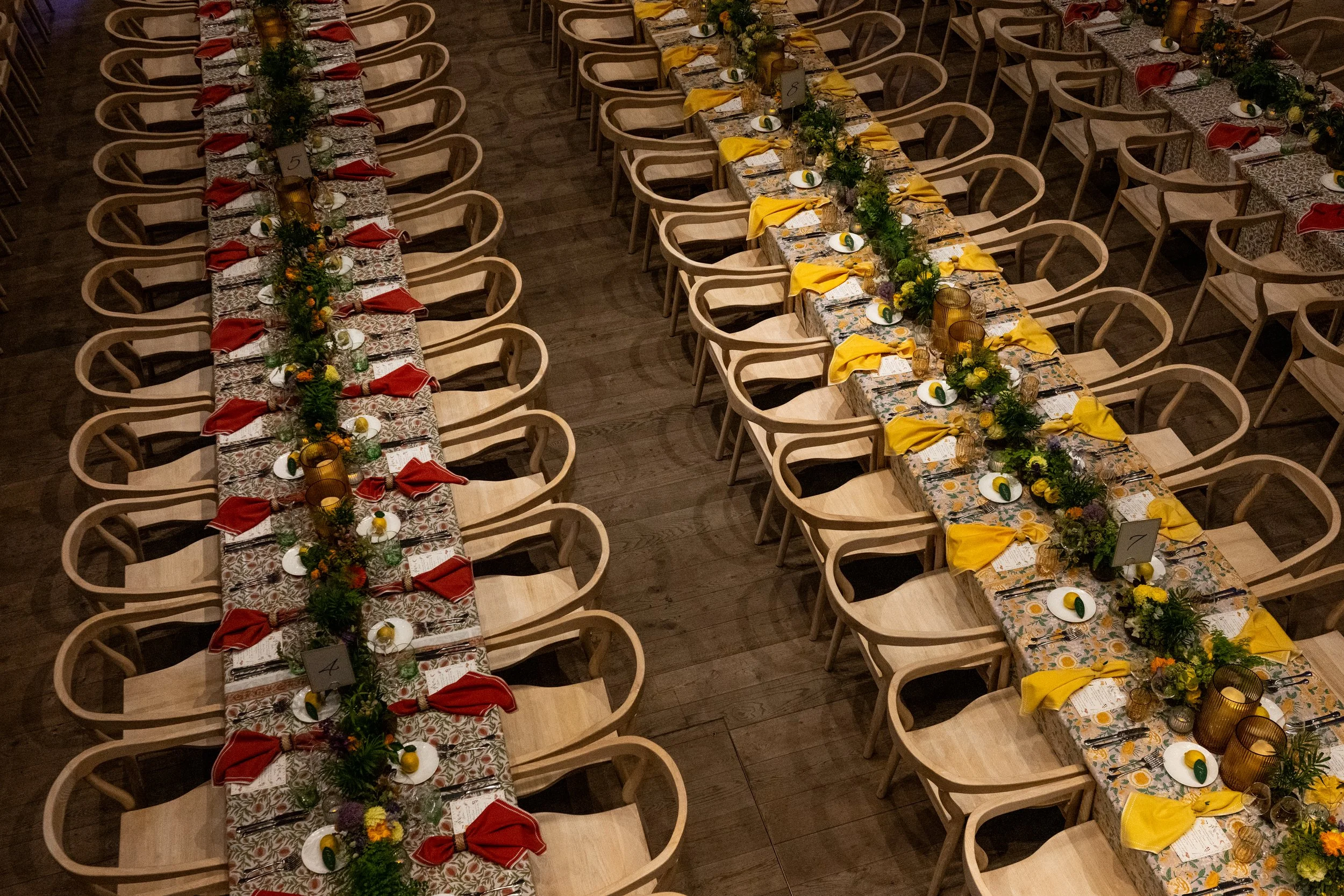 Long banquet tables set for a formal event with colorful floral centerpieces, each place setting includes a floral plate, cutlery, a yellow or red cloth napkin, and decorative glassware, with chairs arranged on either side of the tables.