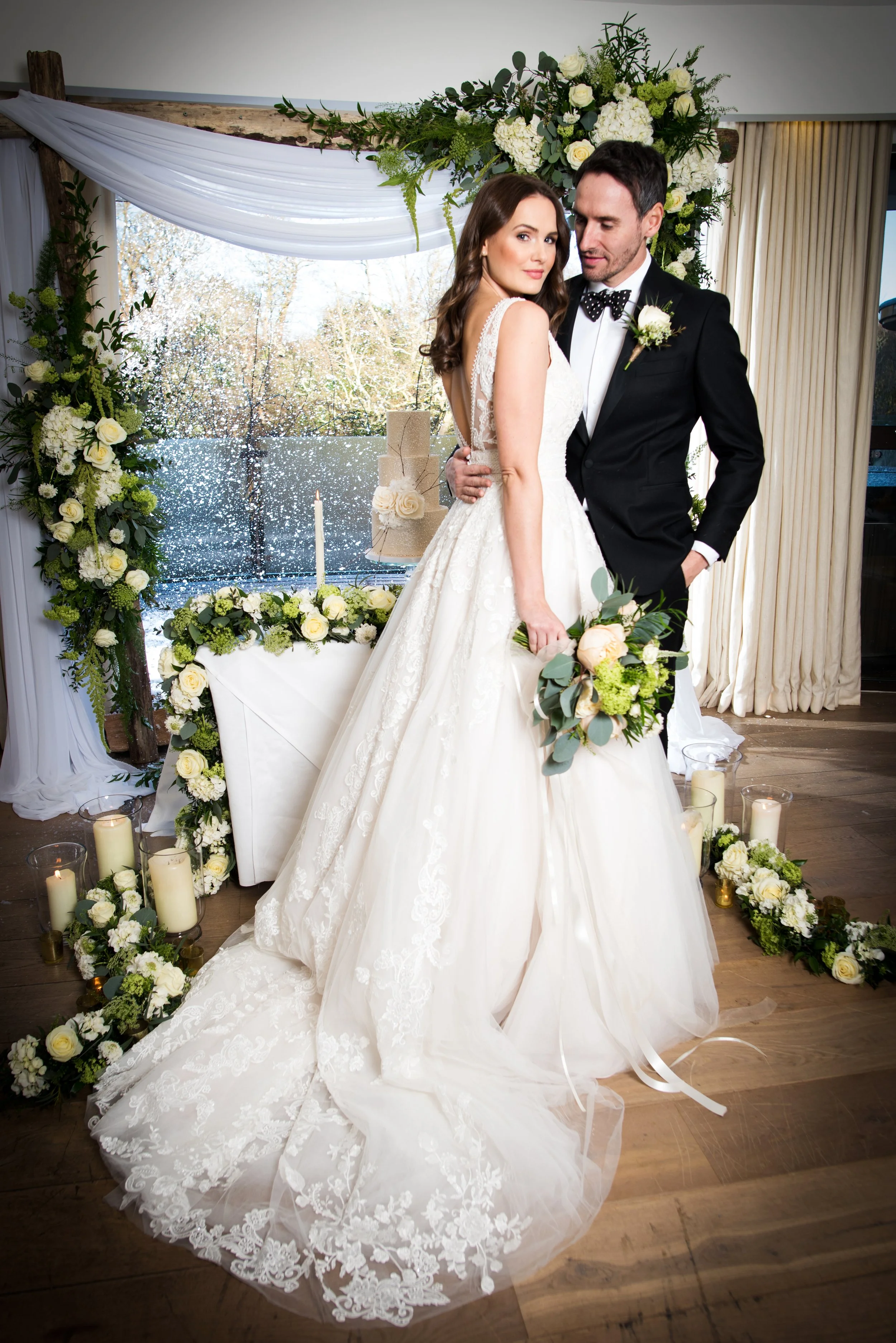 A bride and groom pose for a wedding photo indoors, decorated with white and green floral arrangements, candles, and draped fabric. The bride wears a lace wedding gown, holding a bouquet, and the groom is in a black tuxedo with a bow tie and boutonni