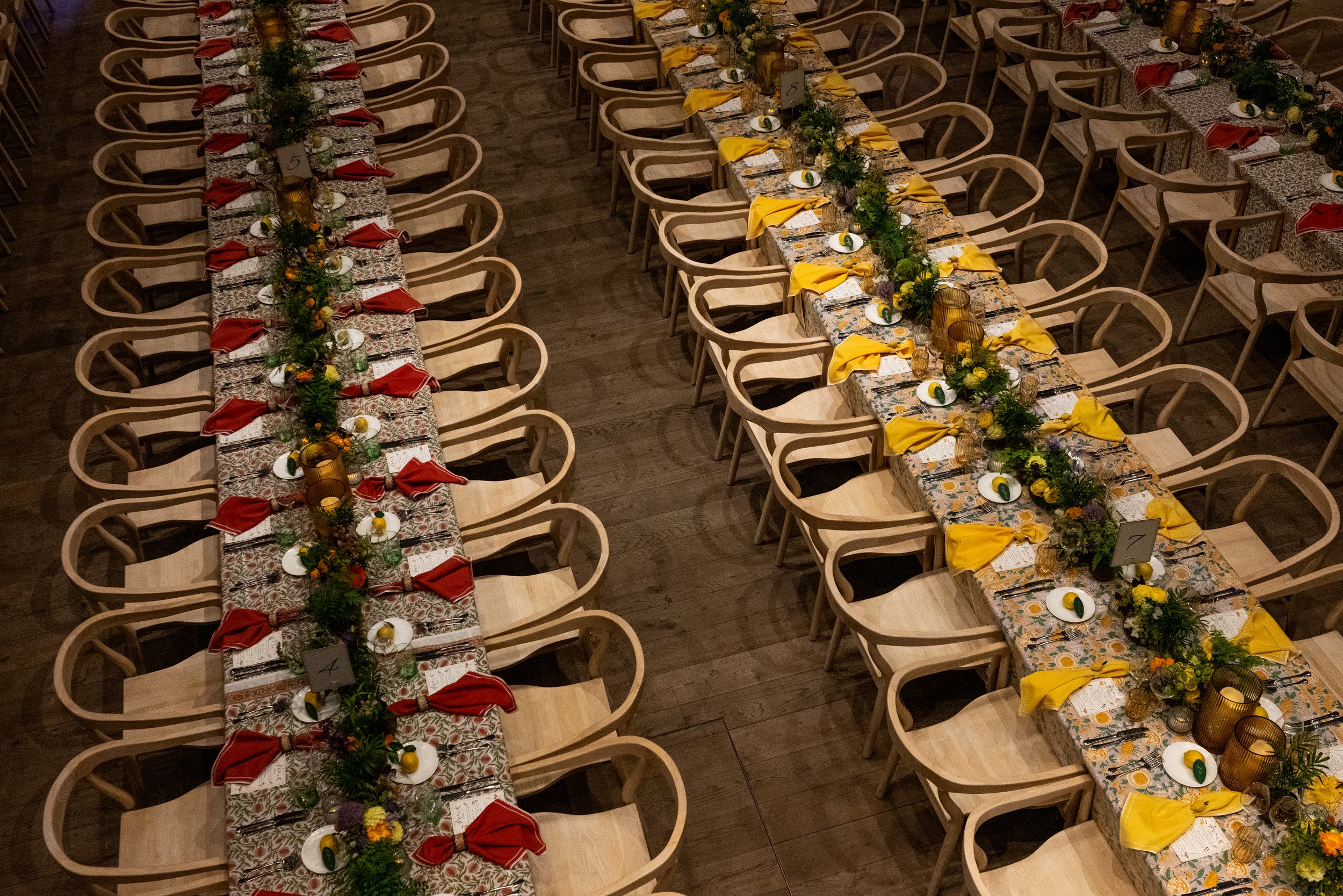 Long banquet tables decorated with colorful tablecloths, greenery and floral centerpieces, yellow and red napkins, glassware, and place settings in an indoor venue with wooden floors.