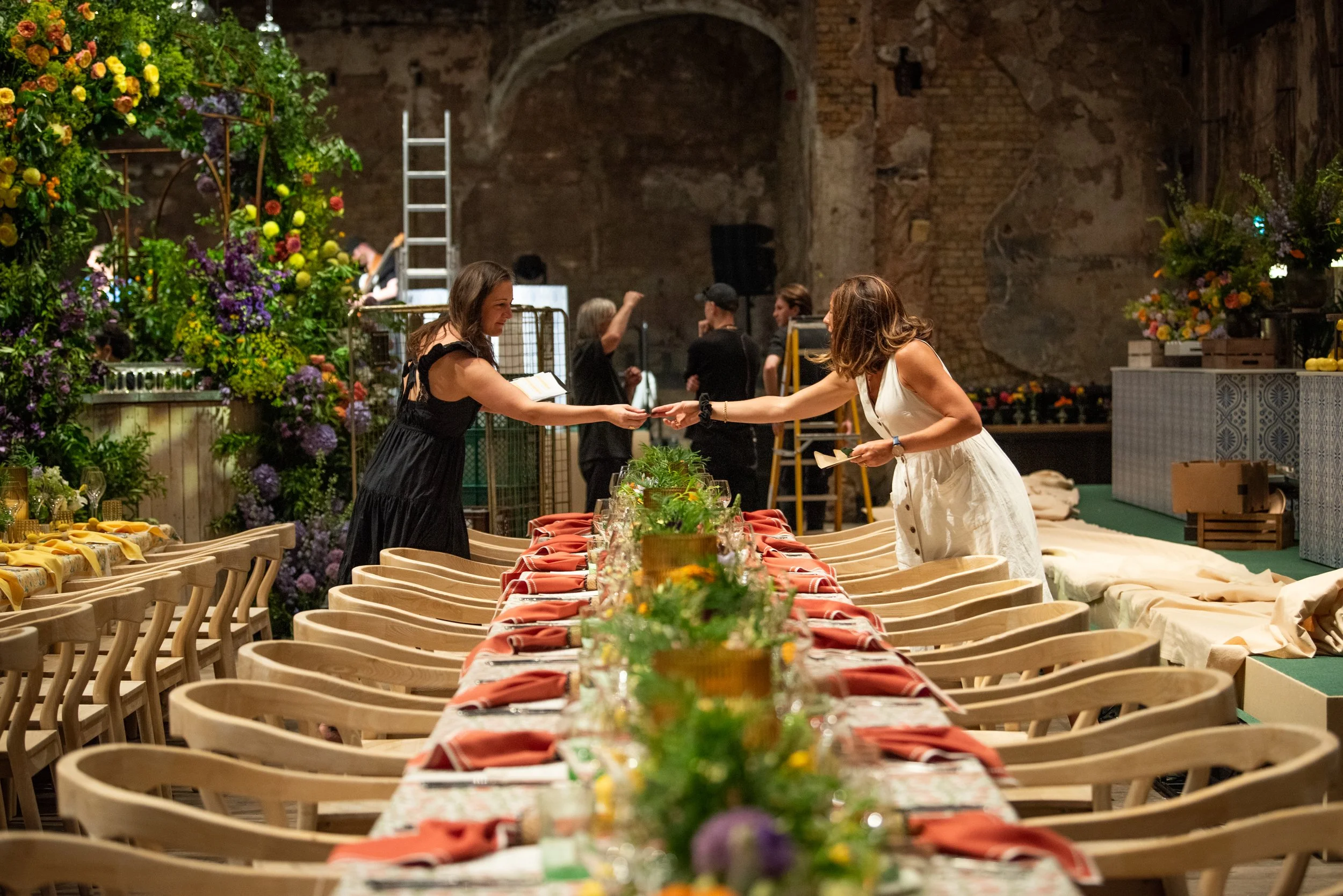 Two women exchanging a handshake at a decorated dining table in an indoor venue with floral arrangements and a brick wall background.