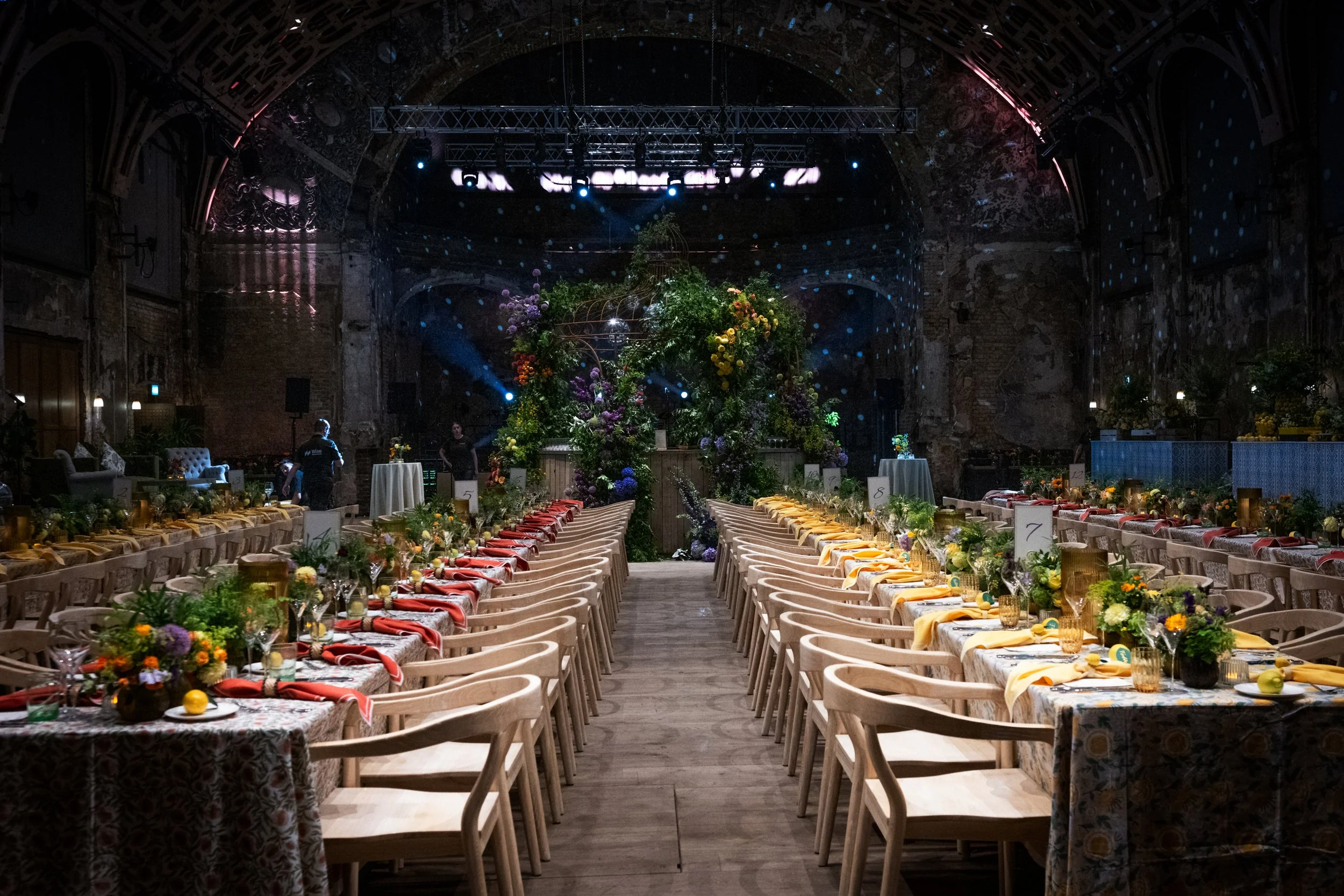Elegant banquet hall with long decorated tables, colorful floral centerpieces, and arranged chairs under a dark, ornate ceiling, prepared for a formal event.