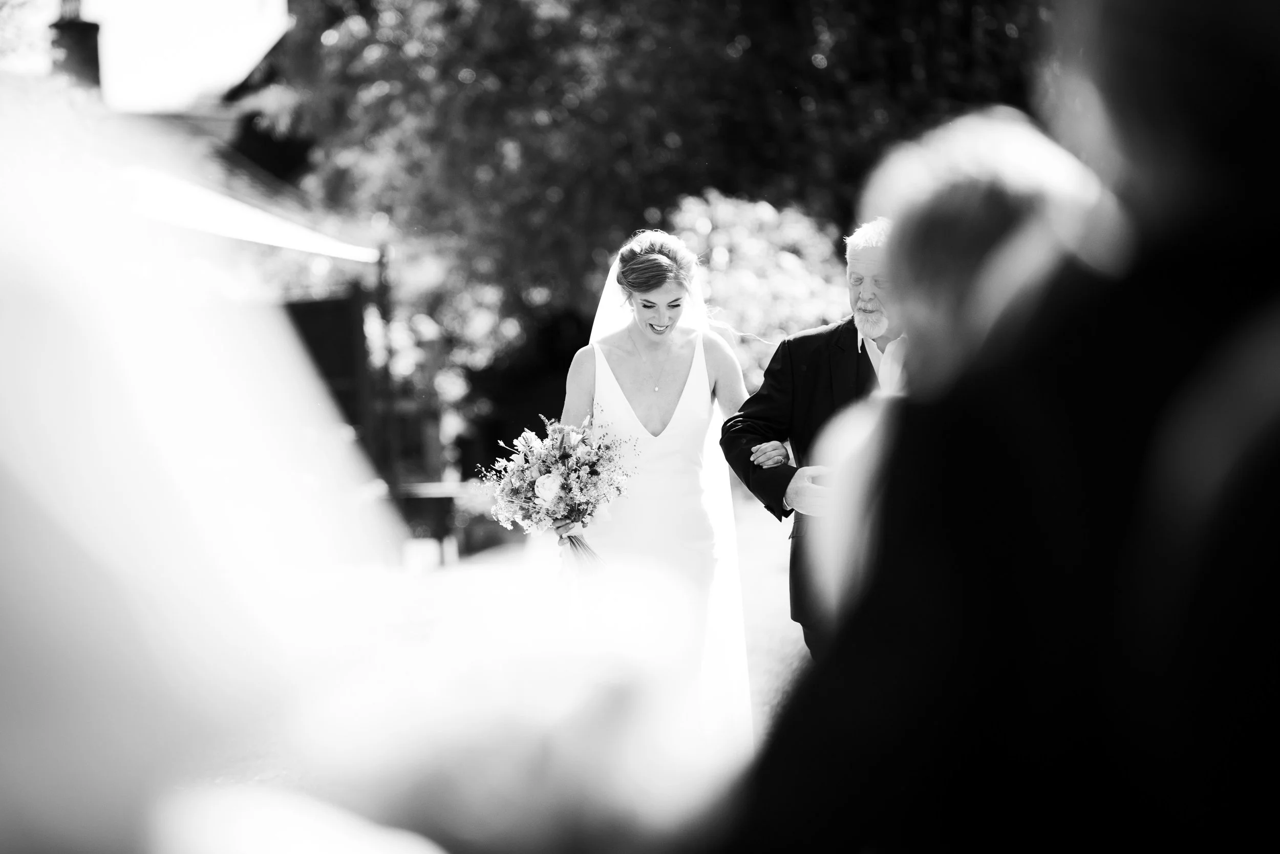 Black and white photo of a bride and older man, likely her father, holding hands at an outdoor wedding ceremony. The bride is smiling and holding a bouquet, and there are blurred figures in the foreground.