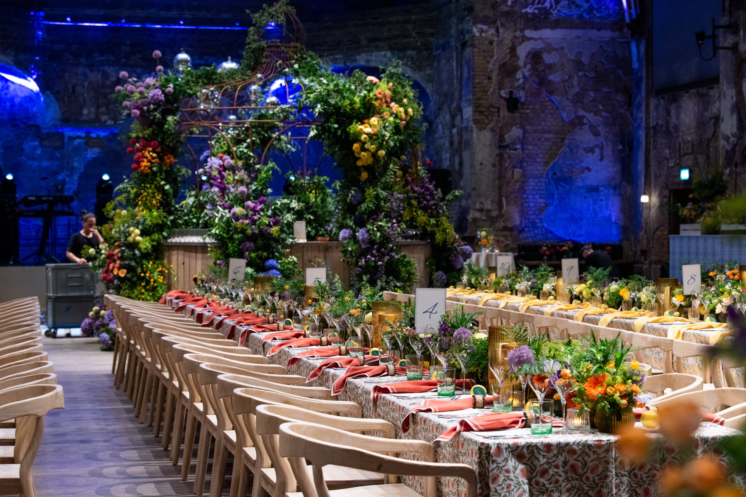 Long banquet table decorated with floral arrangements, tableware, and cloths, set for an event inside an industrial-style venue with exposed brick and stone walls illuminated with blue lighting.