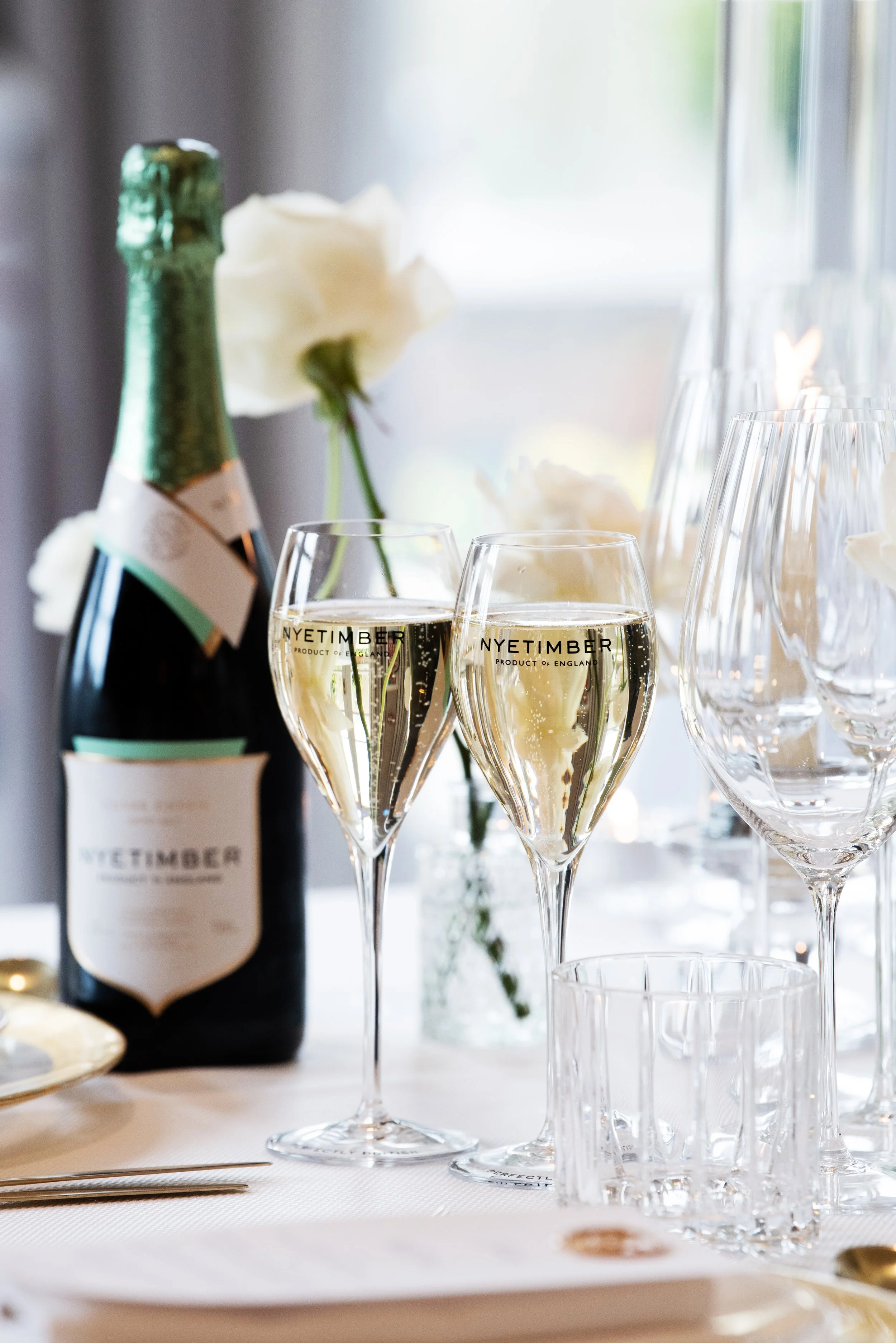 A bottle of Nyetimber sparkling wine, two glasses filled with champagne, and several empty glasses on a table with white tablecloth and flowers in the background.