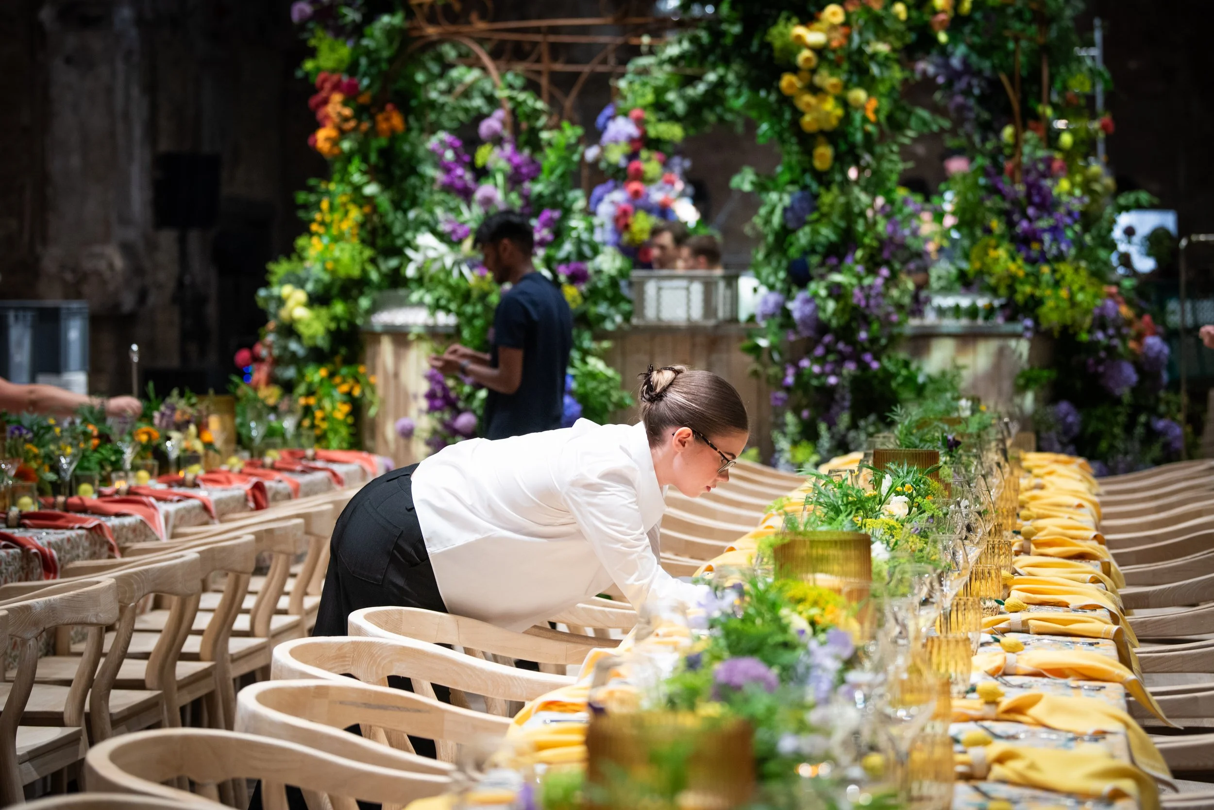 A woman in a white shirt and black pants arranging table settings for a large event with floral arrangements and yellow placemats in a decorated indoor venue.