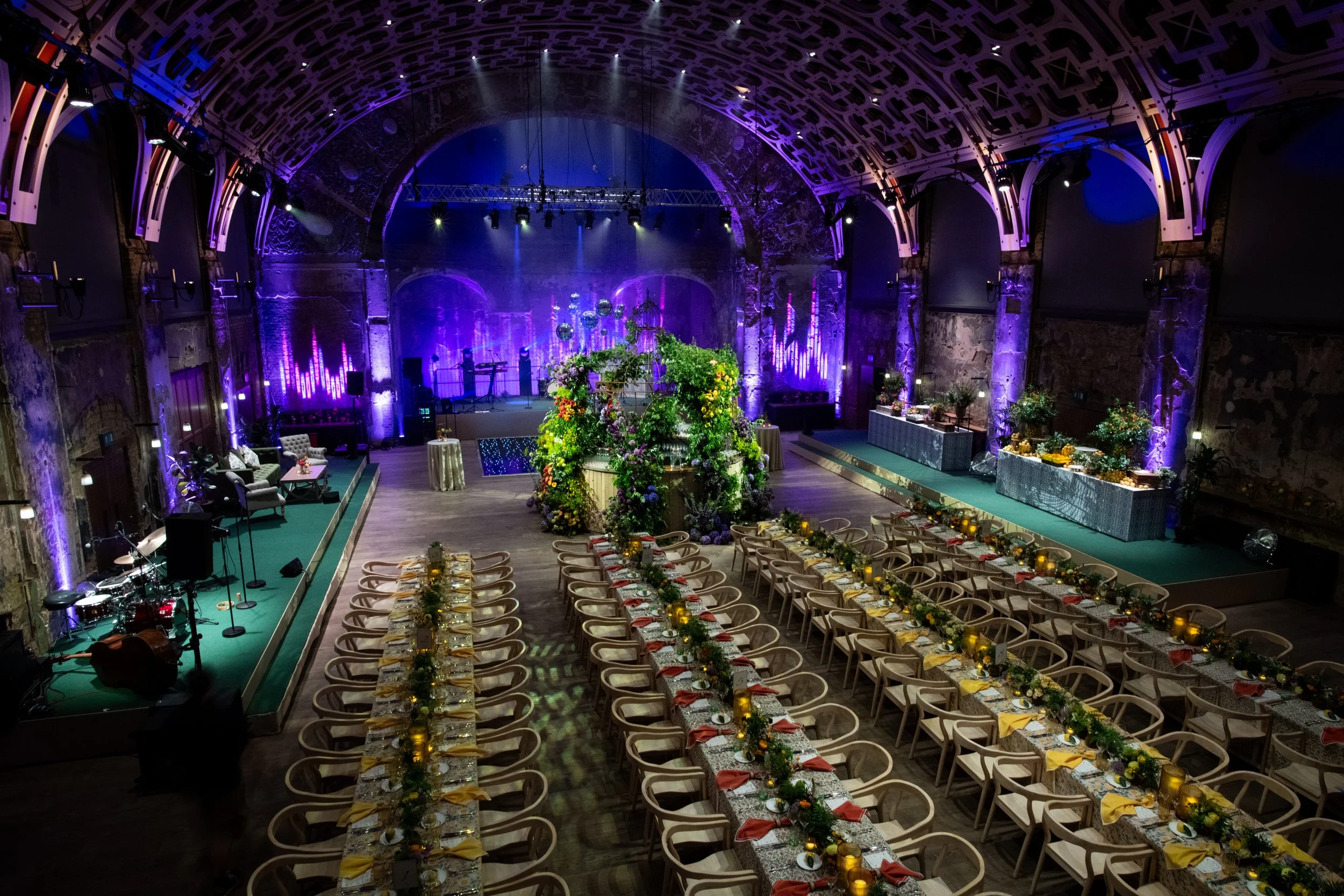 Wedding reception hall decorated with floral arrangements and candlelit tables, a stage with musical instruments, and colorful lighting in a historic stone building with a high arched ceiling.