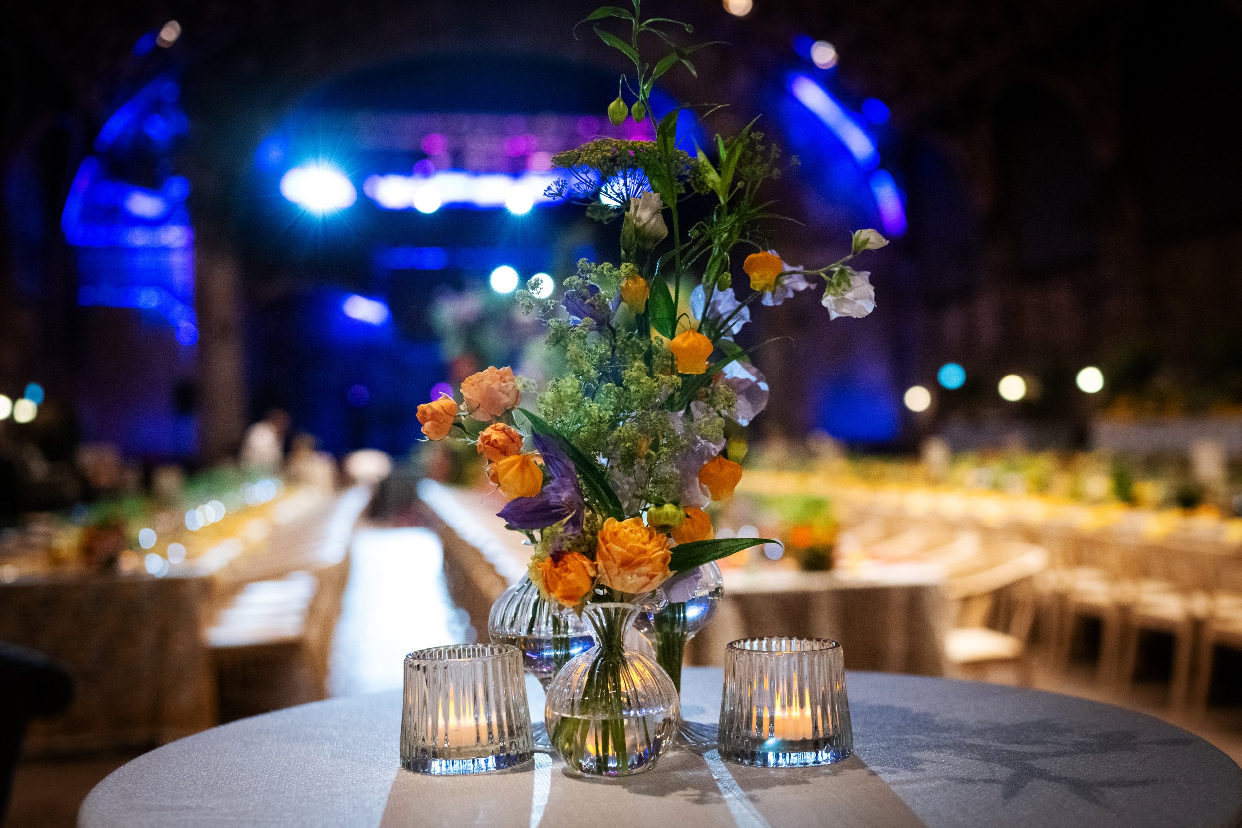A floral centerpiece with various colorful flowers in a glass vase, surrounded by three small glass votive candle holders, on a round table covered with a white tablecloth at a nighttime event.
