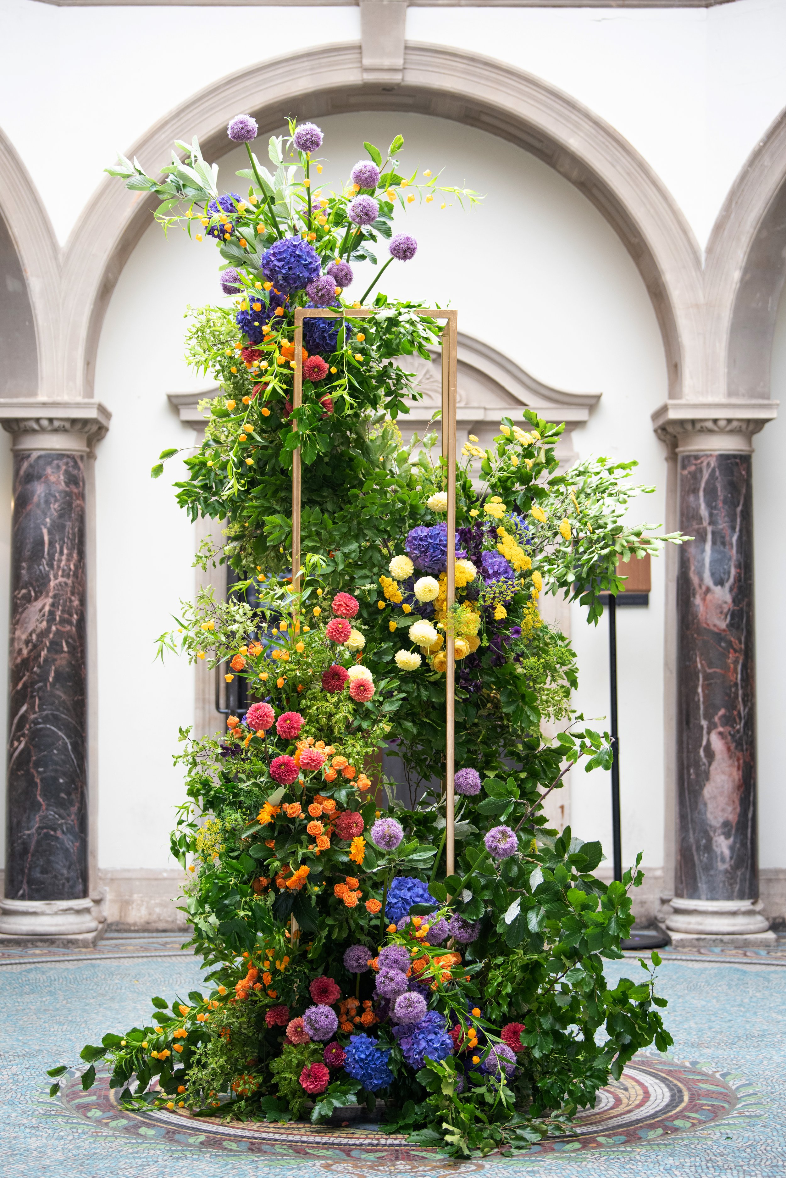 A tall, colorful floral arrangement with various flowers, including purple, orange, yellow, and pink blooms, displayed indoors on a mosaic-tiled floor with marble columns and an arched architectural background.