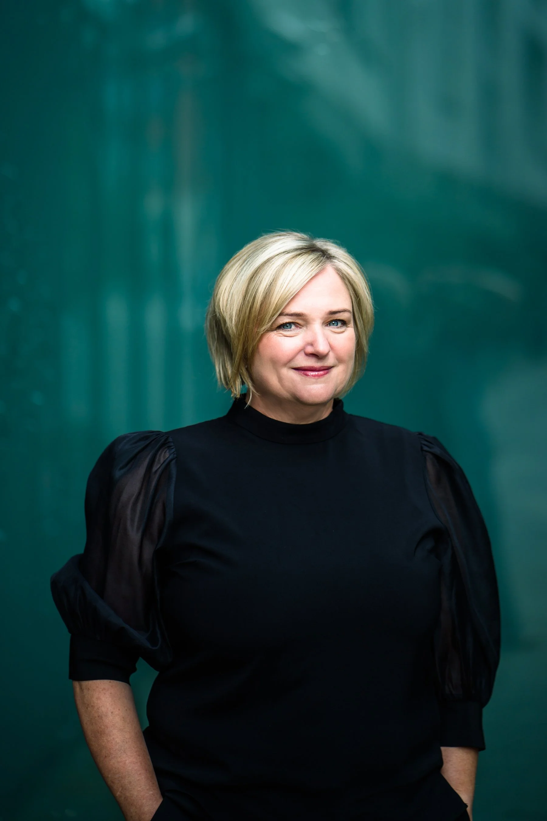 A woman with blonde hair wearing a black top with puffed sleeves standing against a green background.
