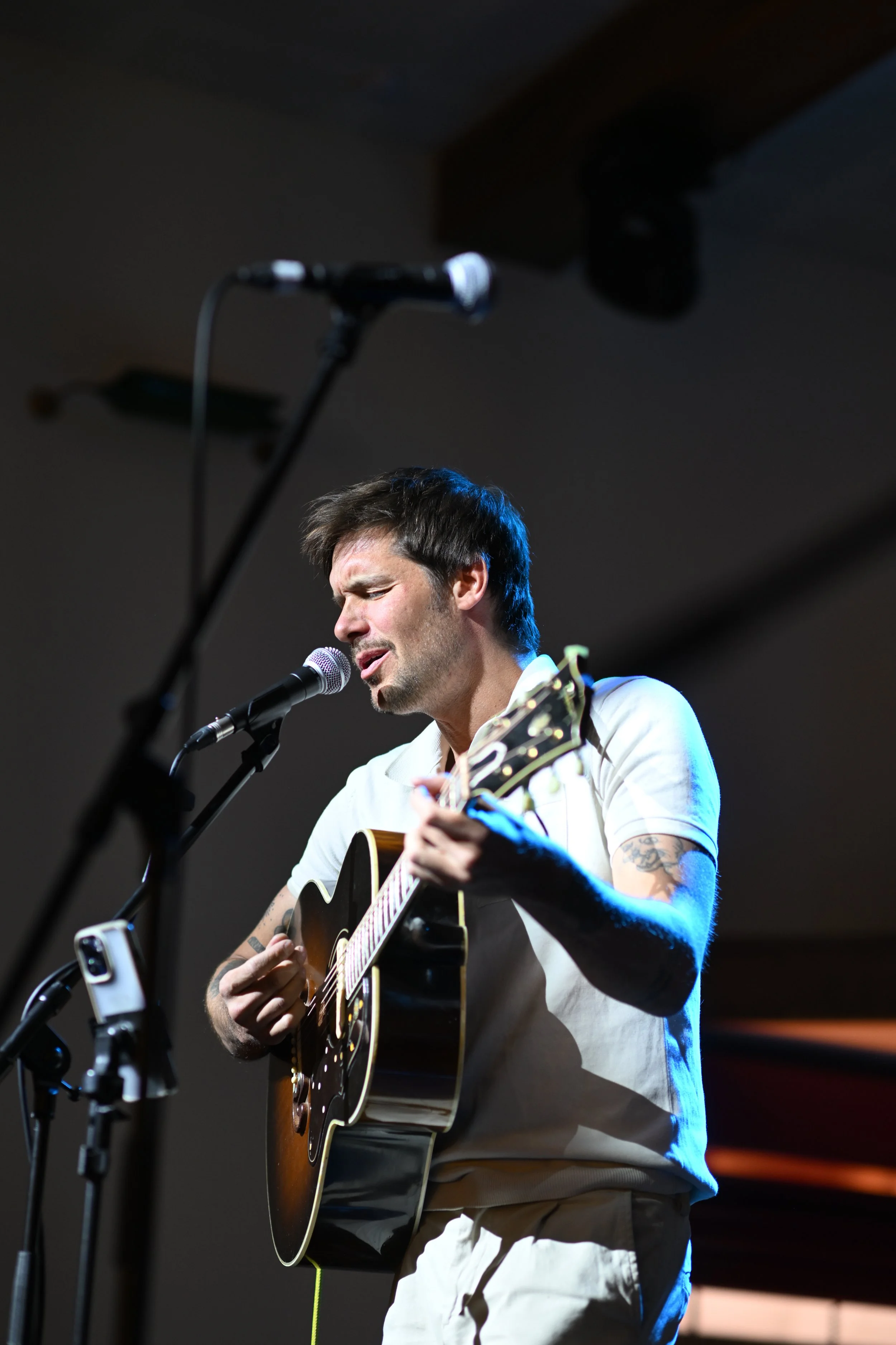 A man with dark hair and tattoos playing an acoustic guitar and singing into a microphone on stage.