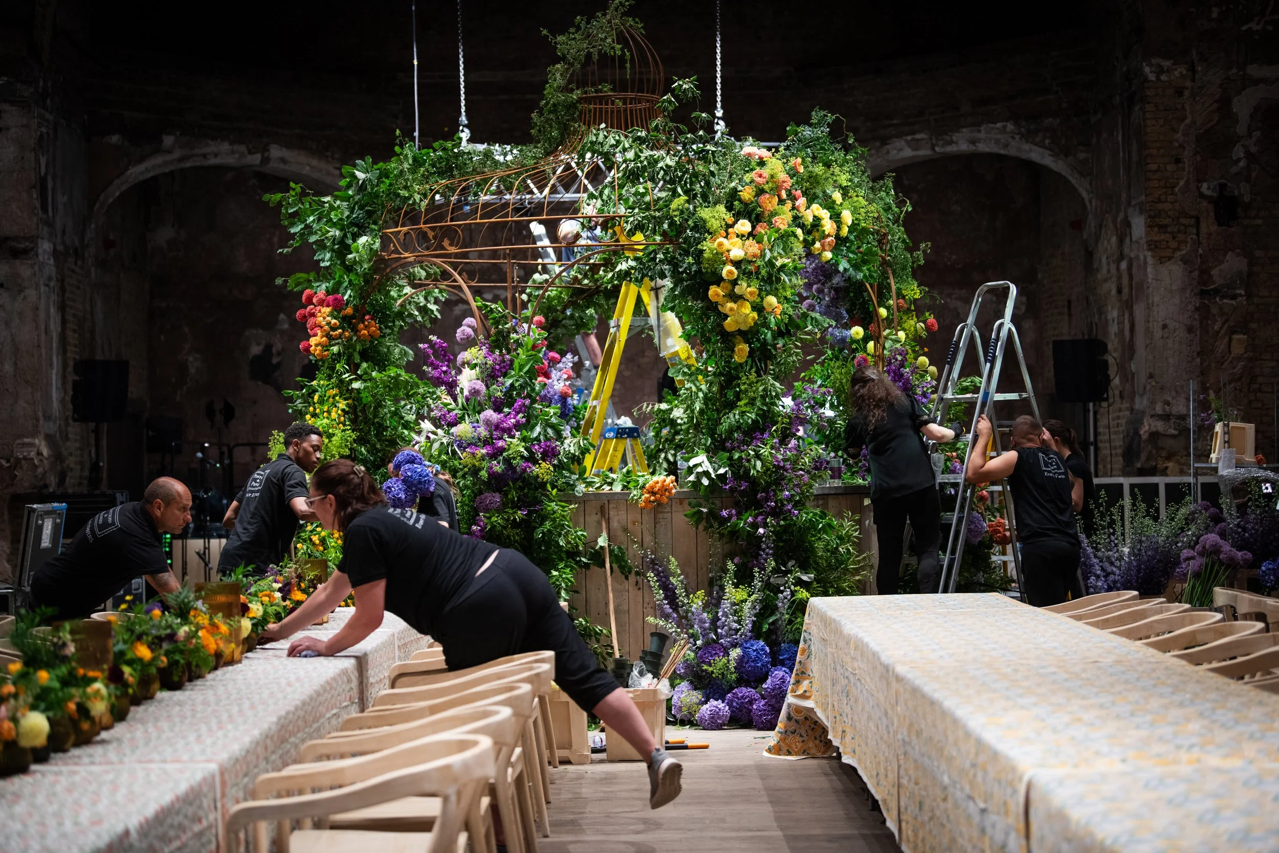 People working together decorating a large floral display indoors, with tables covered in patterned tablecloths and surrounded by chairs, as they arrange and prepare colorful flowers and greenery.