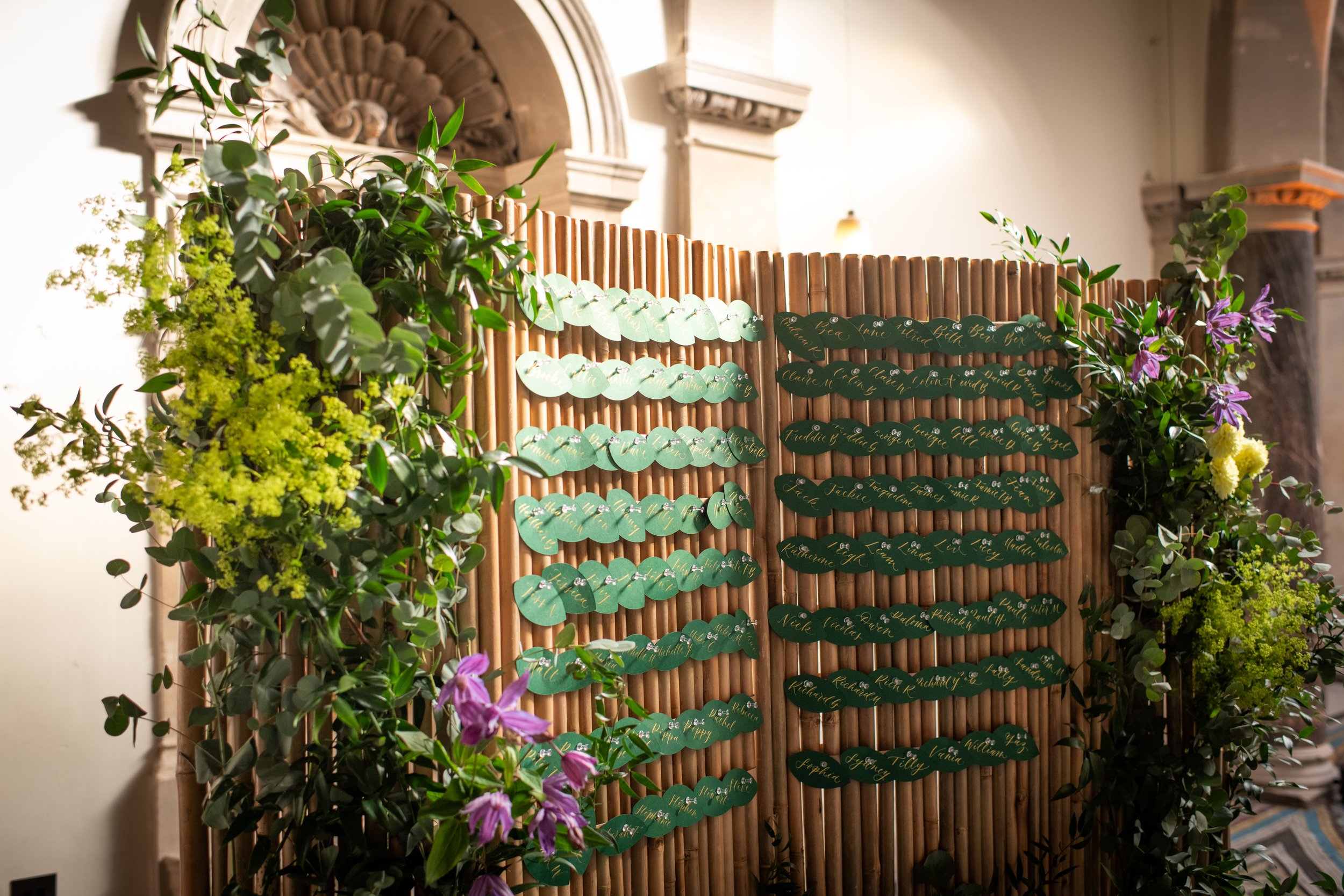 A wooden display board with multiple lines of green paper tags, each with handwritten names in gold ink, surrounded by green foliage and purple flowers, set against a backdrop of classical architectural details.