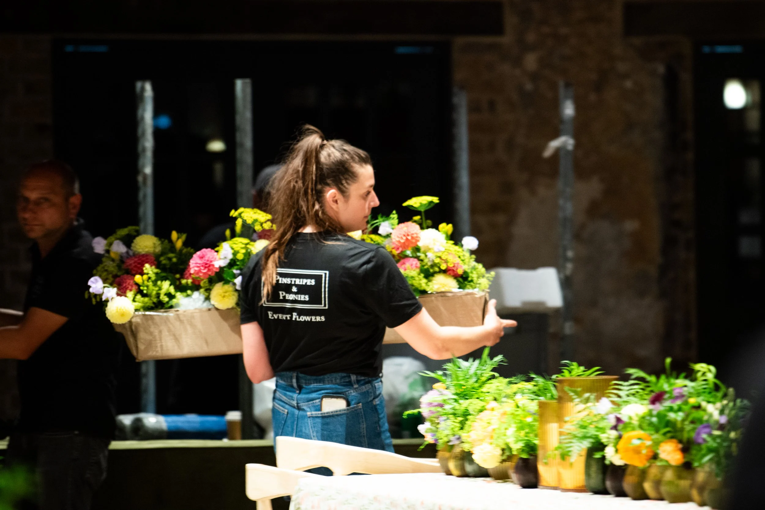 A woman wearing a black T-shirt with white text, holding a large tray or box of colorful flowers at an indoor flower event.