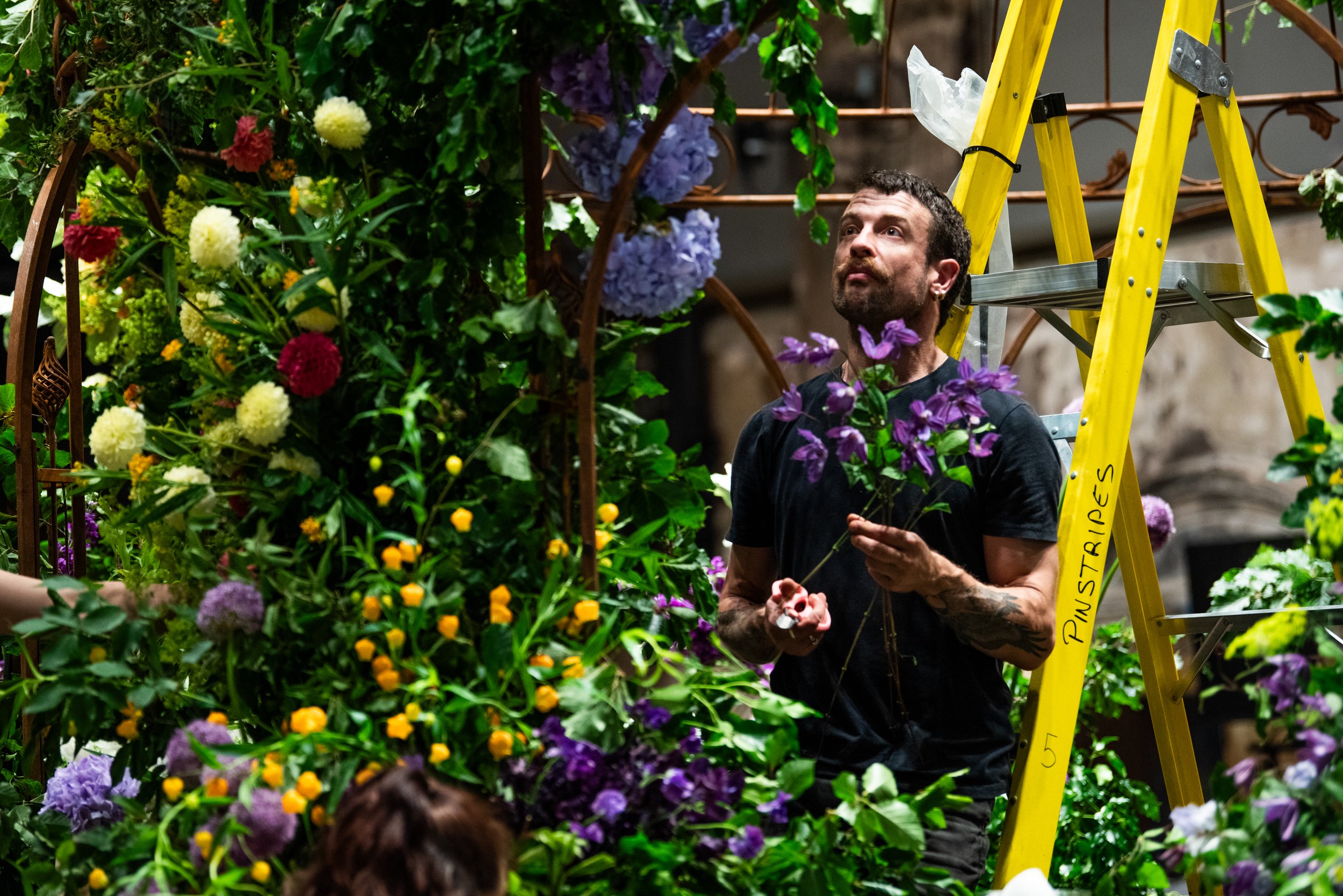 A man with curly hair and tattoos is standing next to a yellow ladder, surrounded by colorful flowers, in what appears to be a floral workshop or garden center.