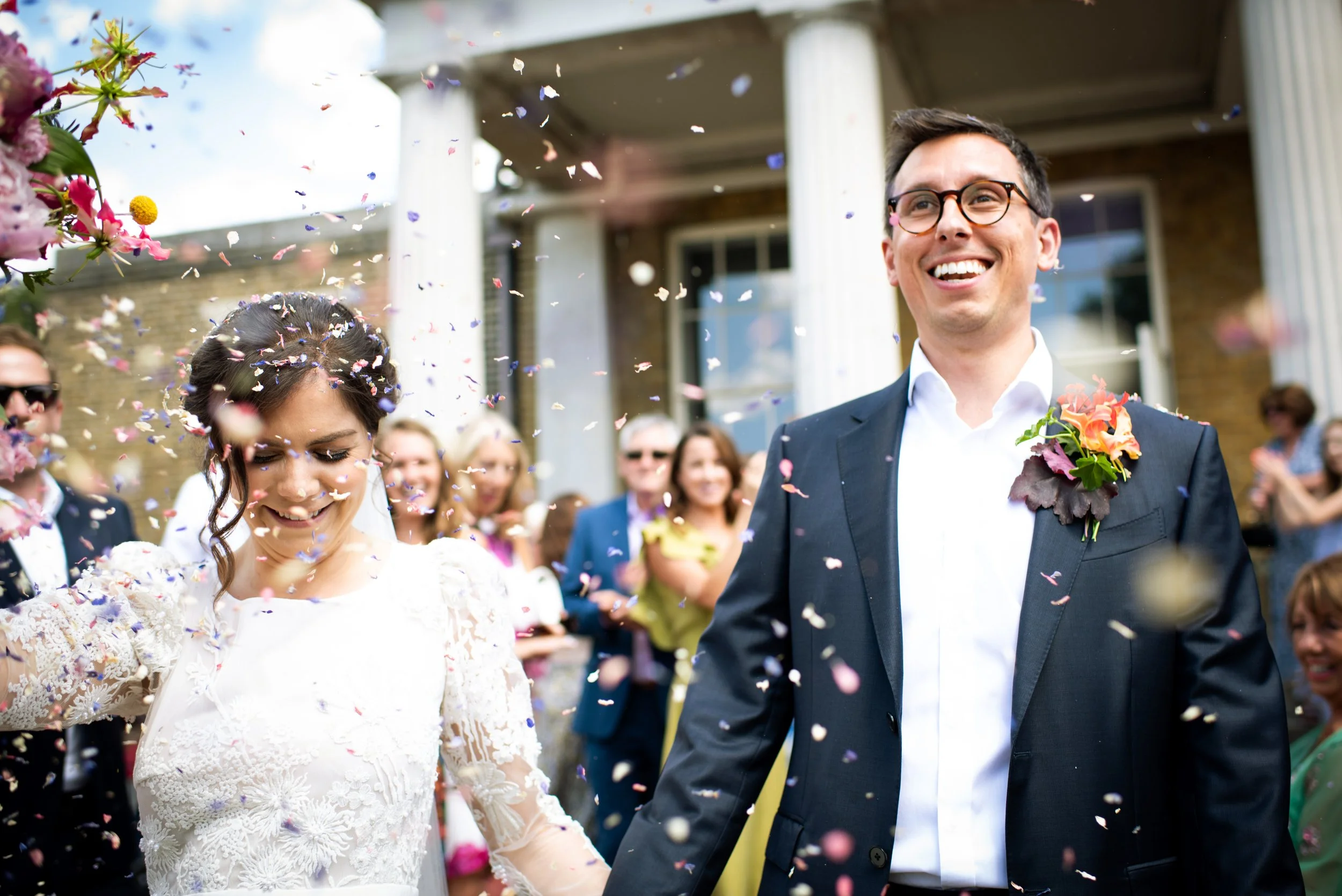 Bride and groom smiling as they walk through confetti on their wedding day, surrounded by happy guests outside a house with large white columns.
