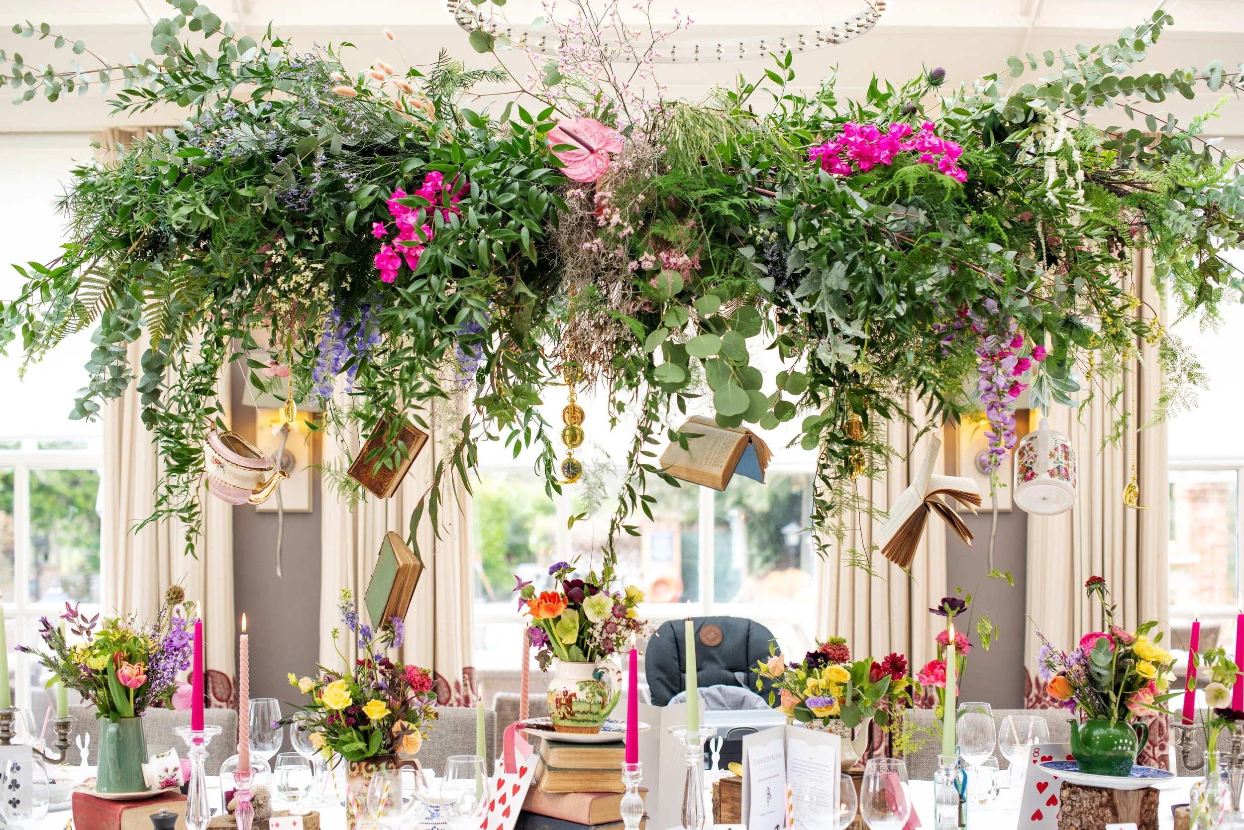 A festive dining table at Chewton Glen decorated with colorful flowers, candles, and hanging ornaments, with a large floral arrangement suspended above, set in a bright room with windows and cream curtains.