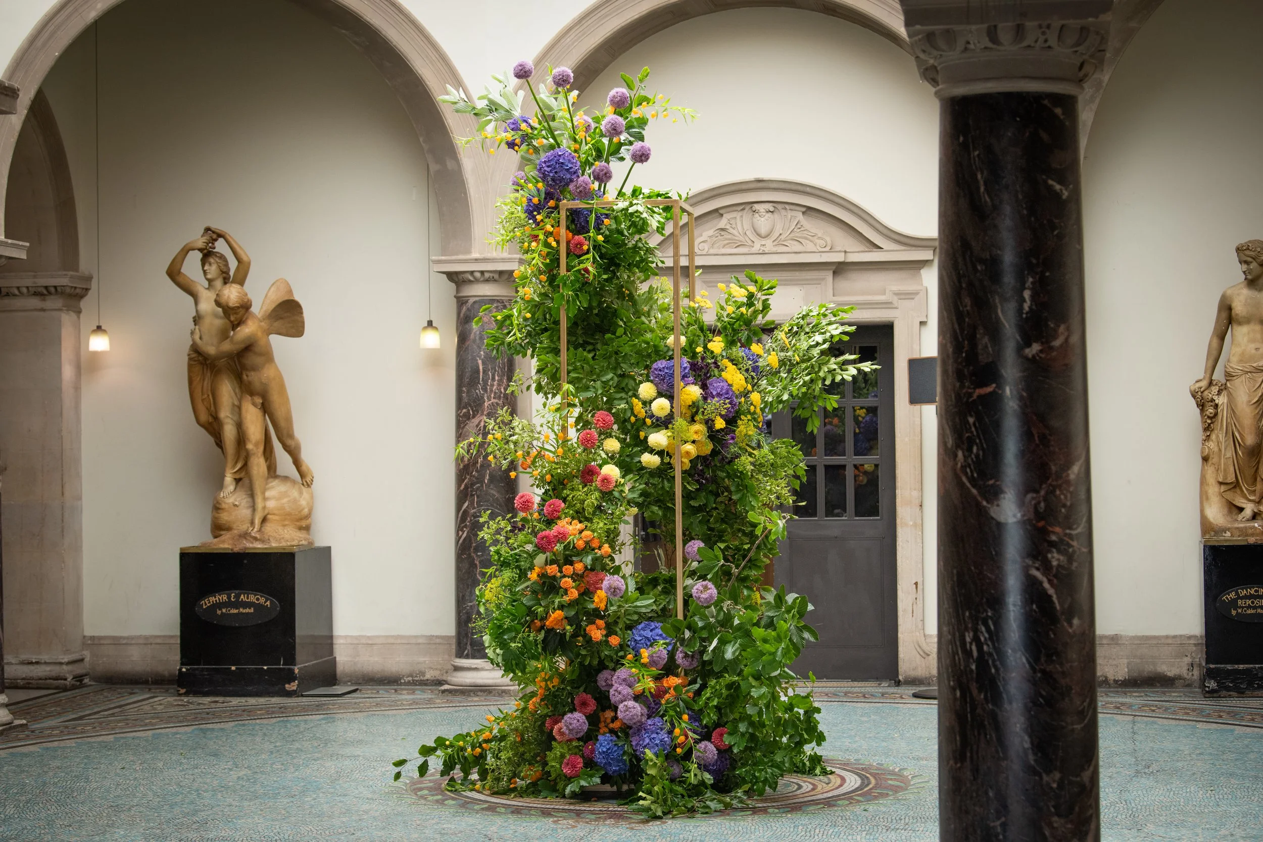 A colorful flower arrangement on a stand in an indoor space with classical architecture, surrounded by marble columns and marble statues.