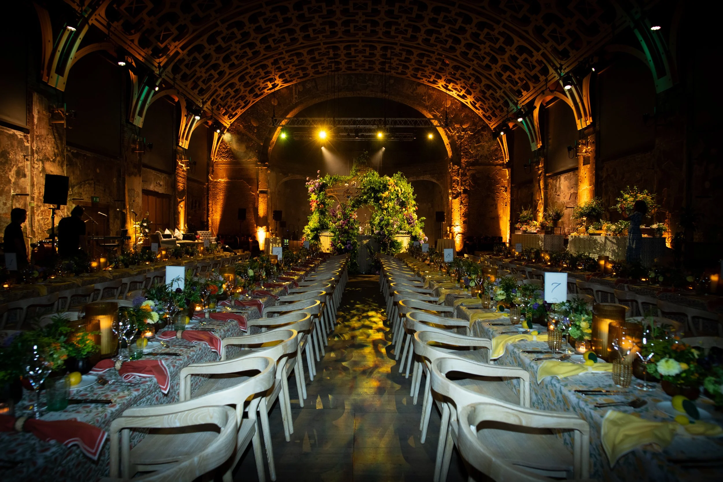 A large indoor event hall with an arched ceiling and exposed brick walls, decorated for a formal gathering with long tables covered in patterned tablecloths, floral centerpieces, candles, and place settings, with soft warm lighting and stage lights i