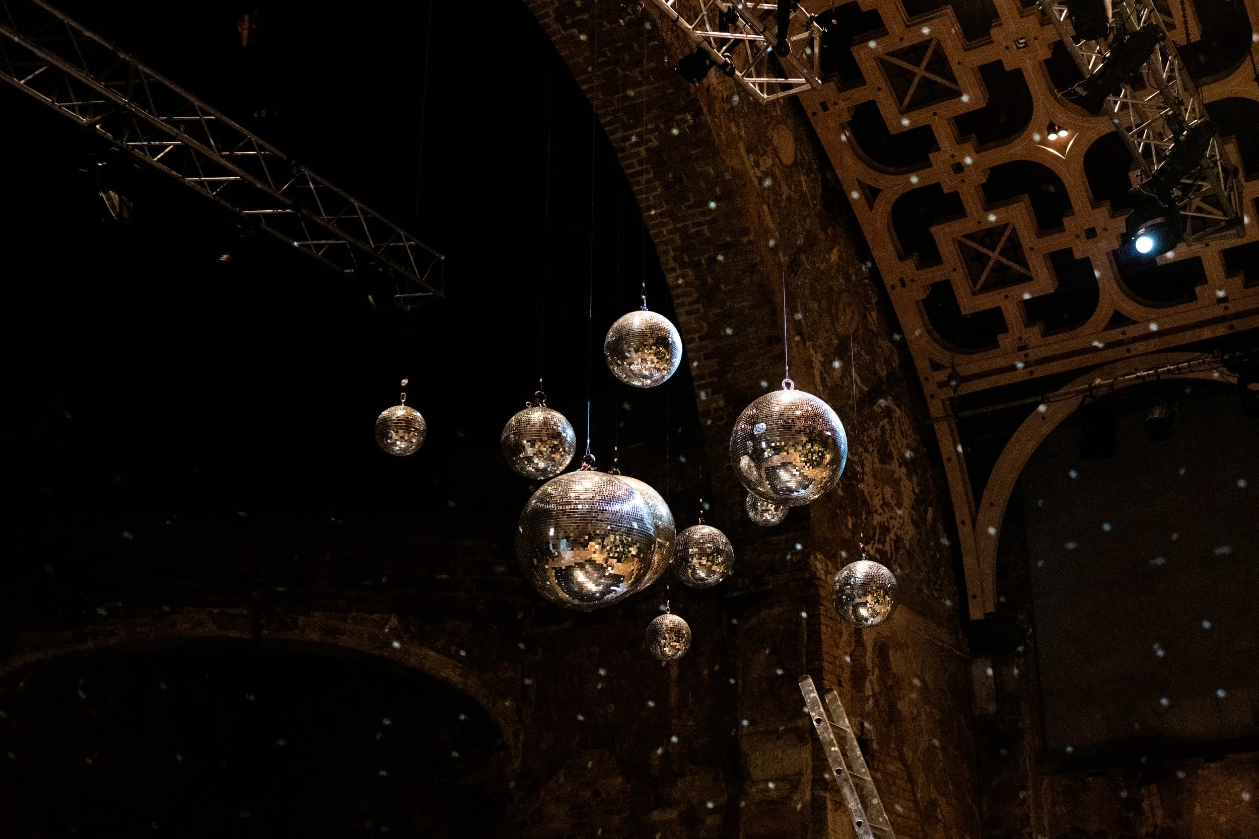 Multiple disco balls hanging from the ceiling of a venue, reflecting light and creating a sparkling effect, with stage lighting equipment and wooden architectural elements visible.