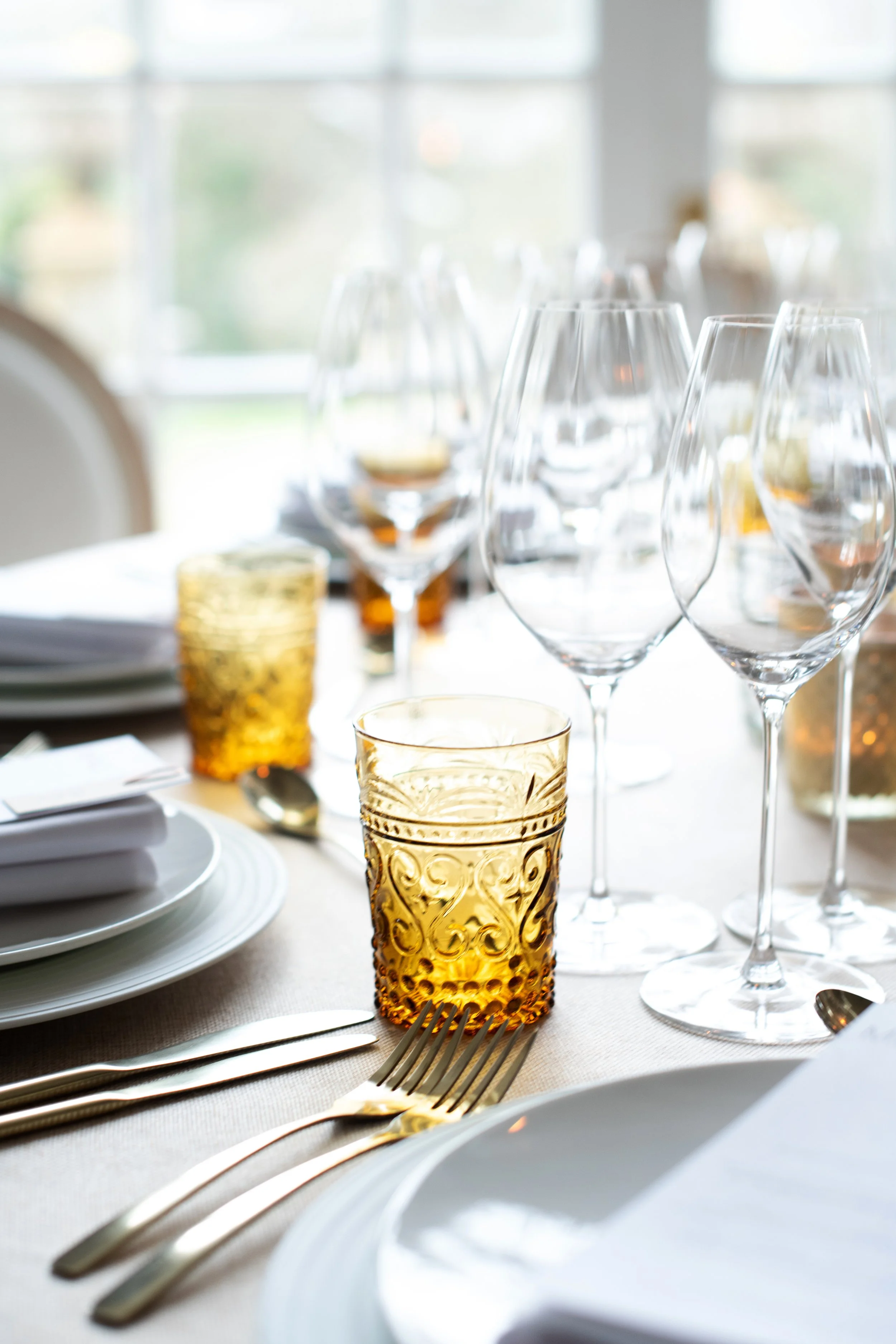 Elegant table setting with clear wine glasses and amber-colored textured glass tumblers, white plates, and silverware on a beige tablecloth, illuminated by natural light.