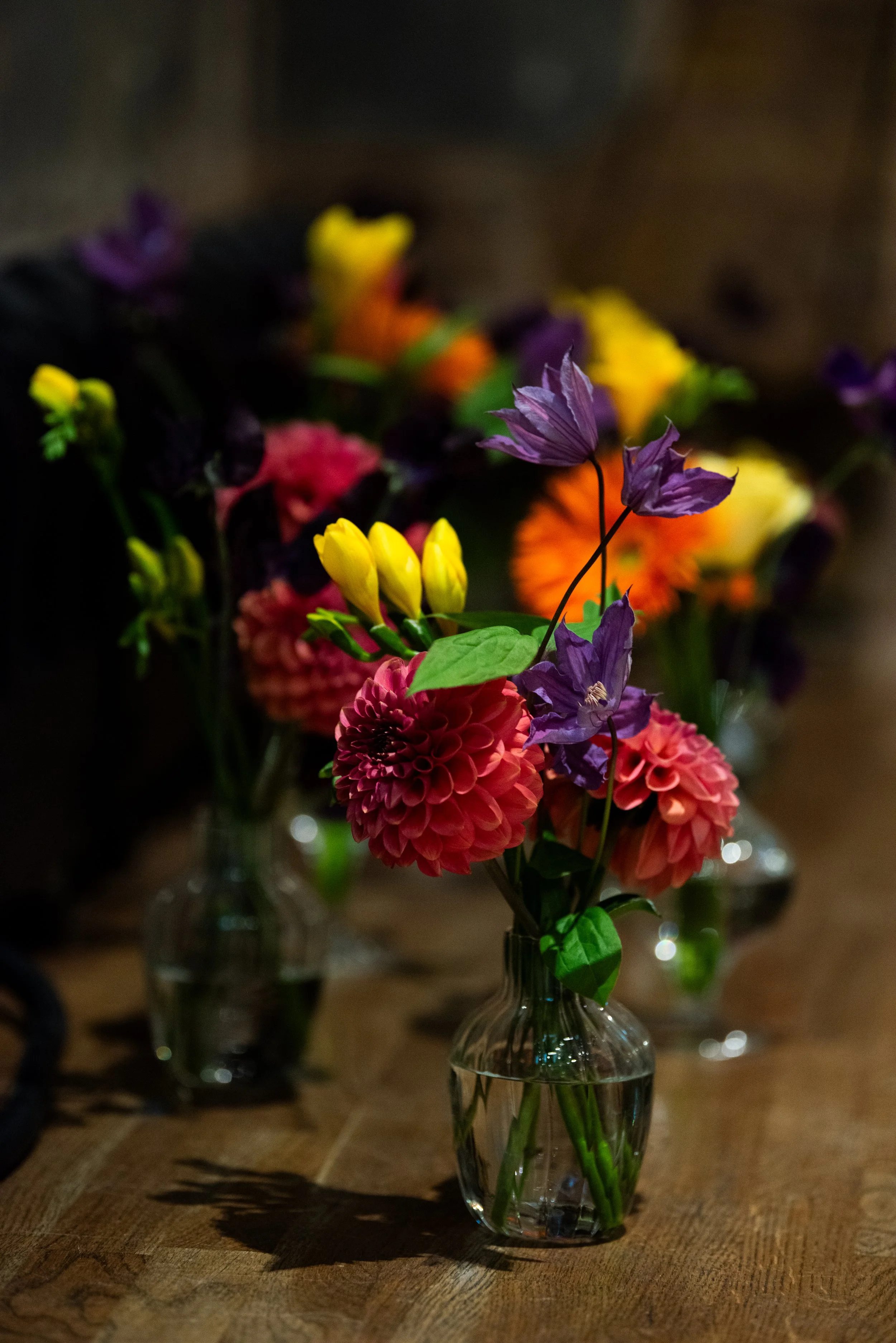 A glass vase with a bouquet of colorful flowers including purple, yellow, orange, and pink, placed on a wooden surface with a blurred background.