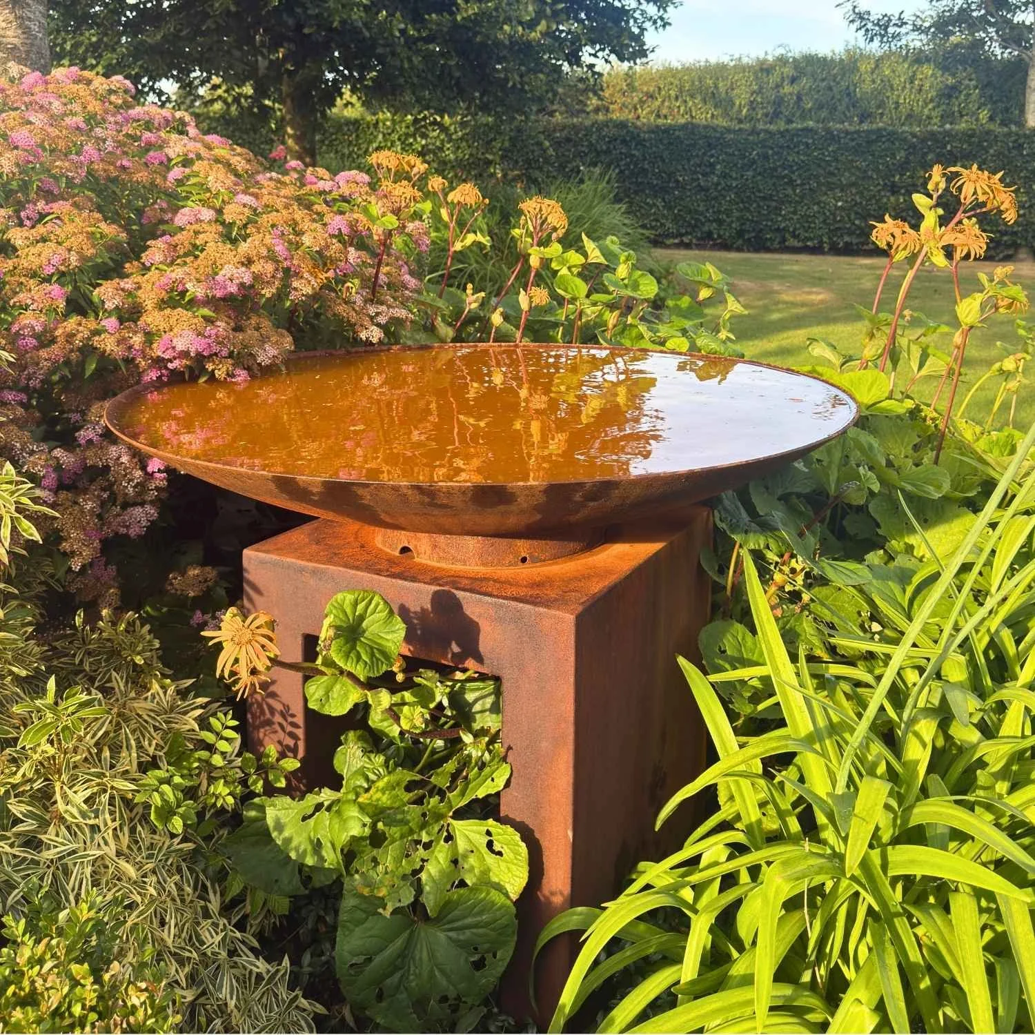 Rusted corten steel water bowl feature mounted on a square U-shaped pedestal set in a garden.⁠