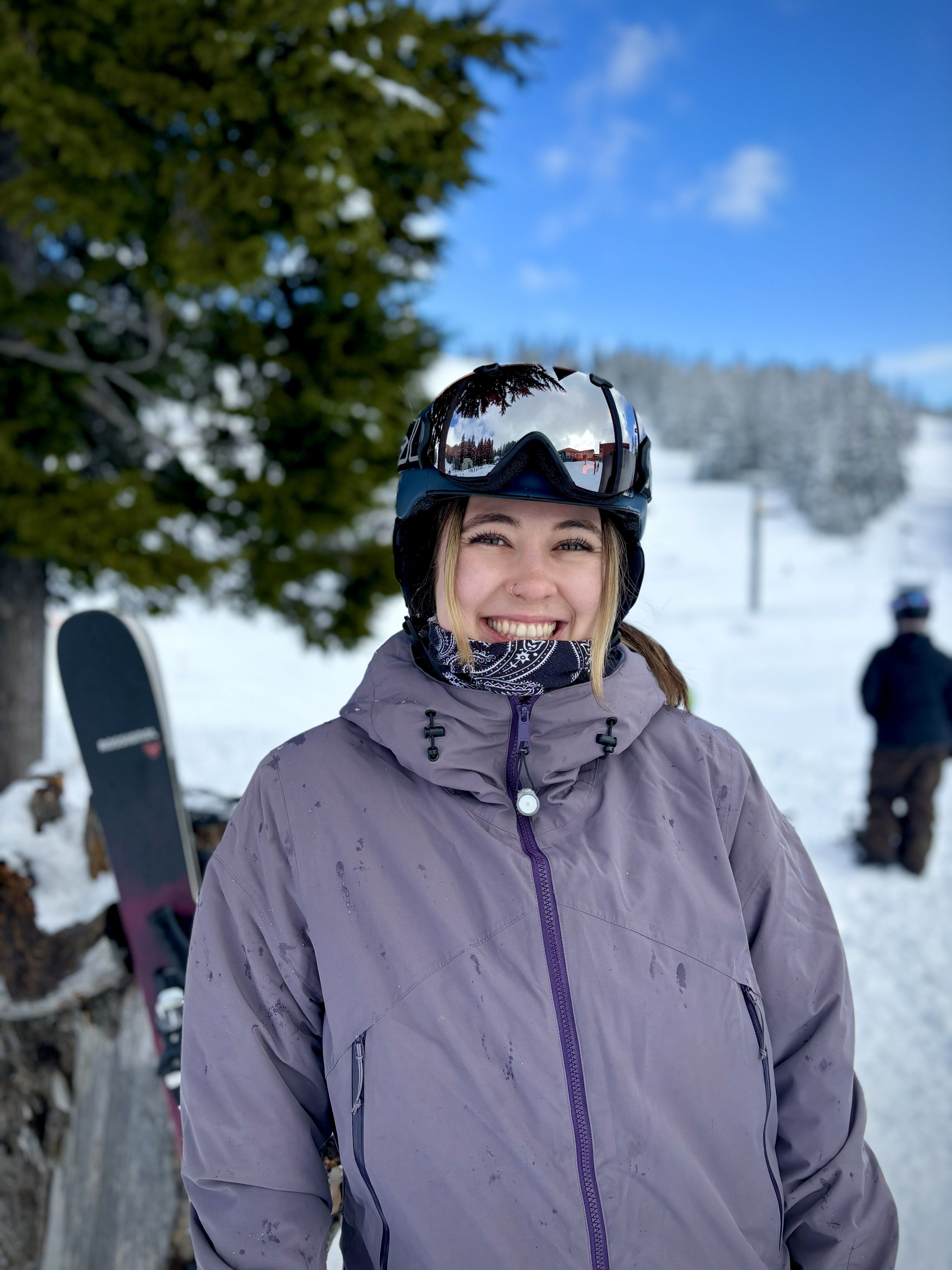 A woman smiling in ski gear outdoors on a snowy mountain, with a purple jacket, black ski helmet with goggles, and a neck gaiter. In the background, snow-covered trees and a person in winter clothing.