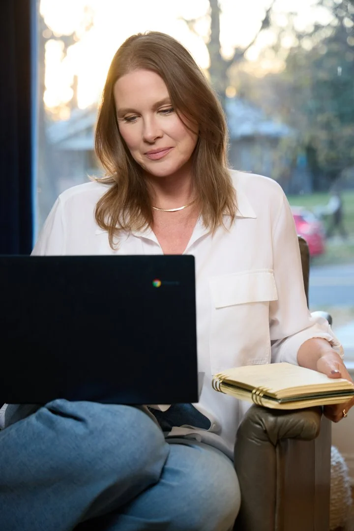 A woman sitting by a window with sunlight in the background, using a laptop and holding a closed notebook.