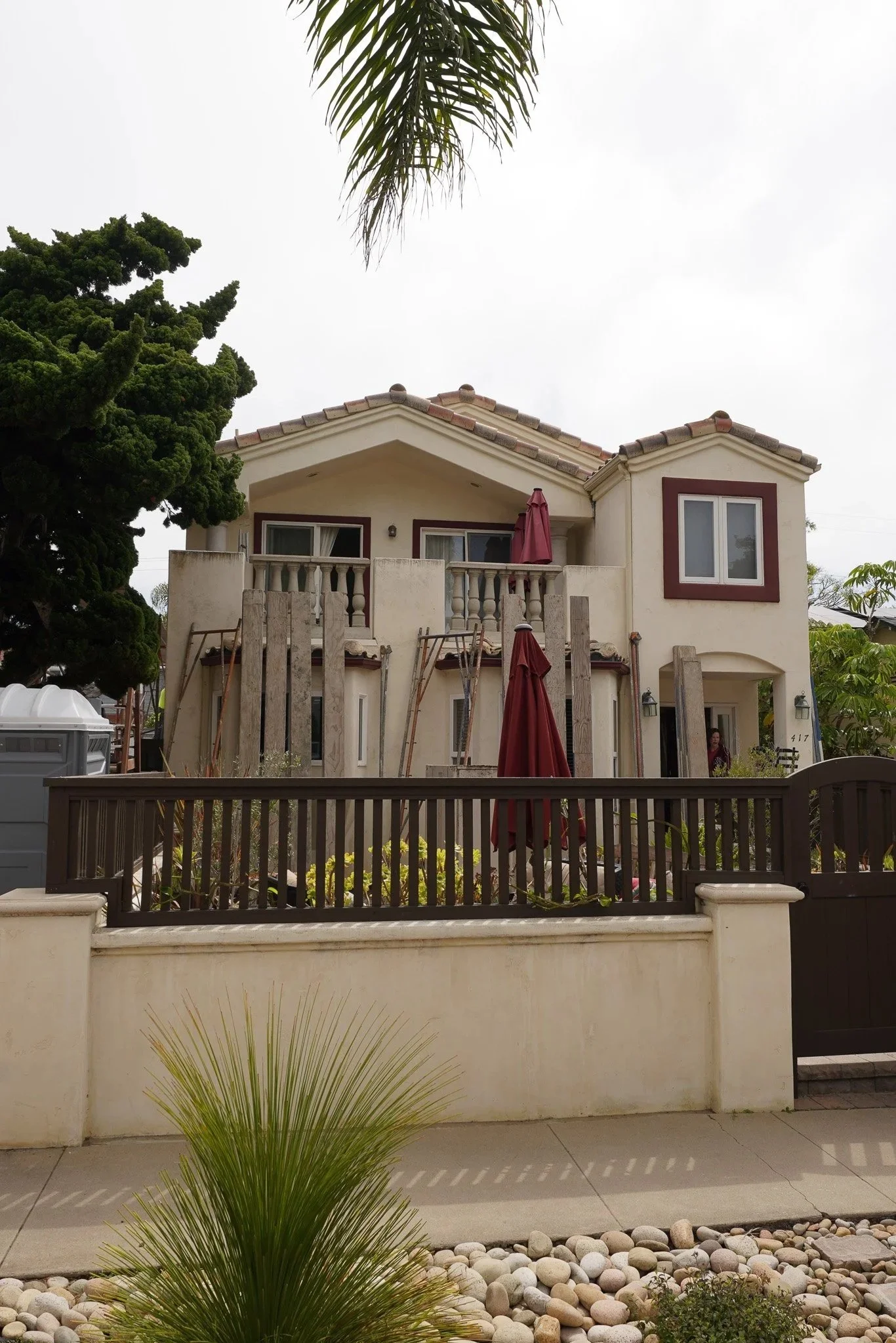 A two-story house with white stucco walls, red trim around the windows, and a terracotta tile roof. The house has a front porch with a small balcony above, partially obscured by a tree and wooden construction materials. A red patio umbrella is on the porch. There is a fence in front, and a landscaped yard with plants, rocks, and a tall palm tree