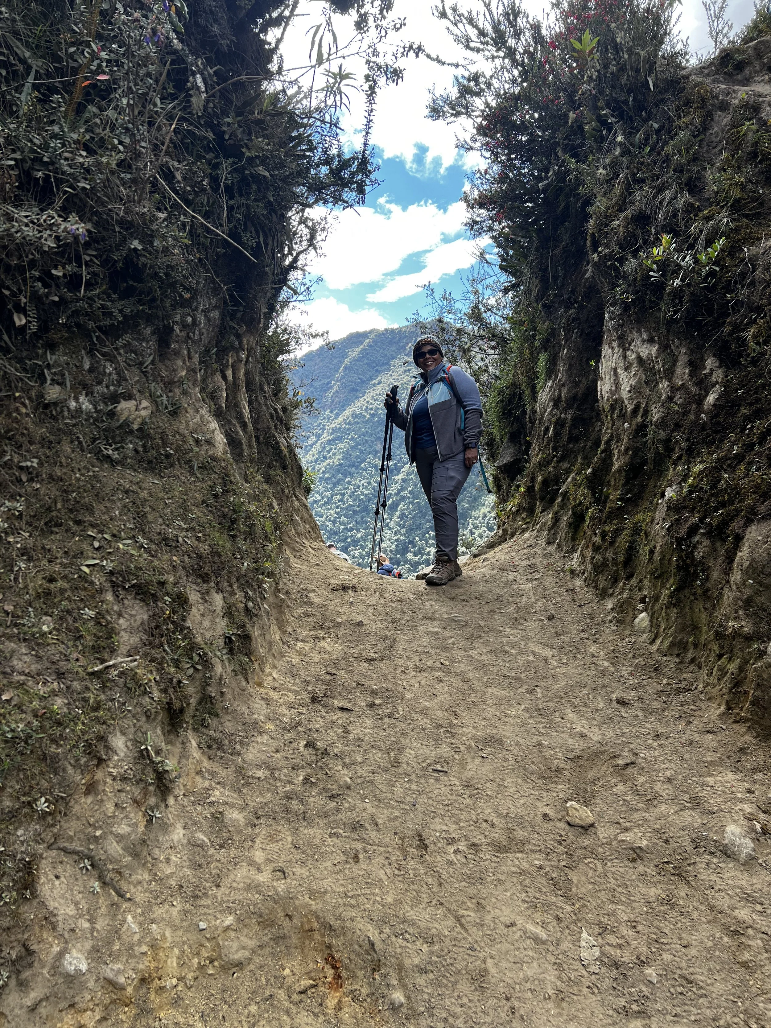 A person hiking on a dirt trail through a narrow canyon or pass with steep, rocky walls, surrounded by vegetation and mountains in the background, under a partly cloudy sky.