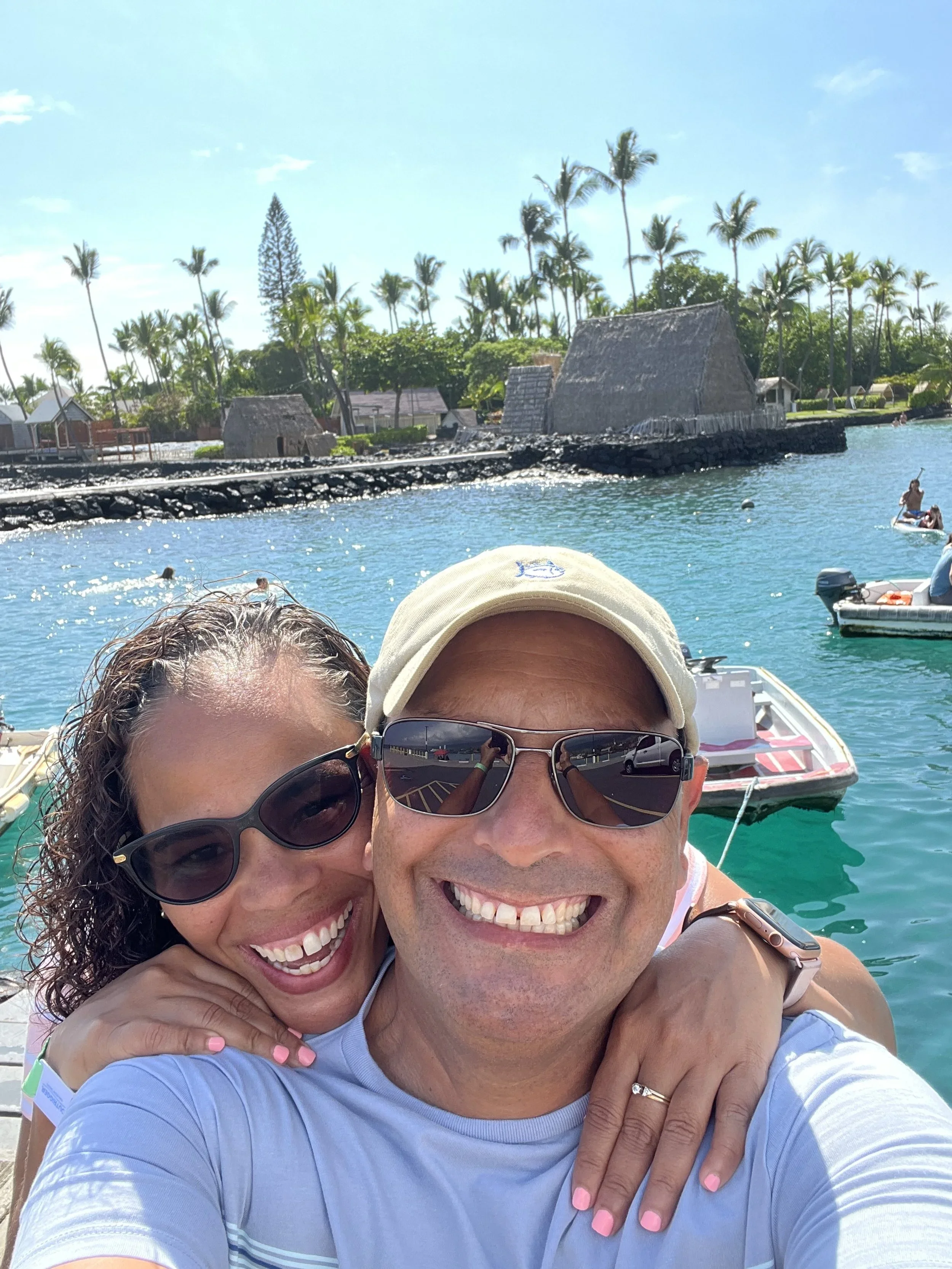 Smiling man and woman taking a selfie on a boat near the water with palm trees and thatched buildings in the background.