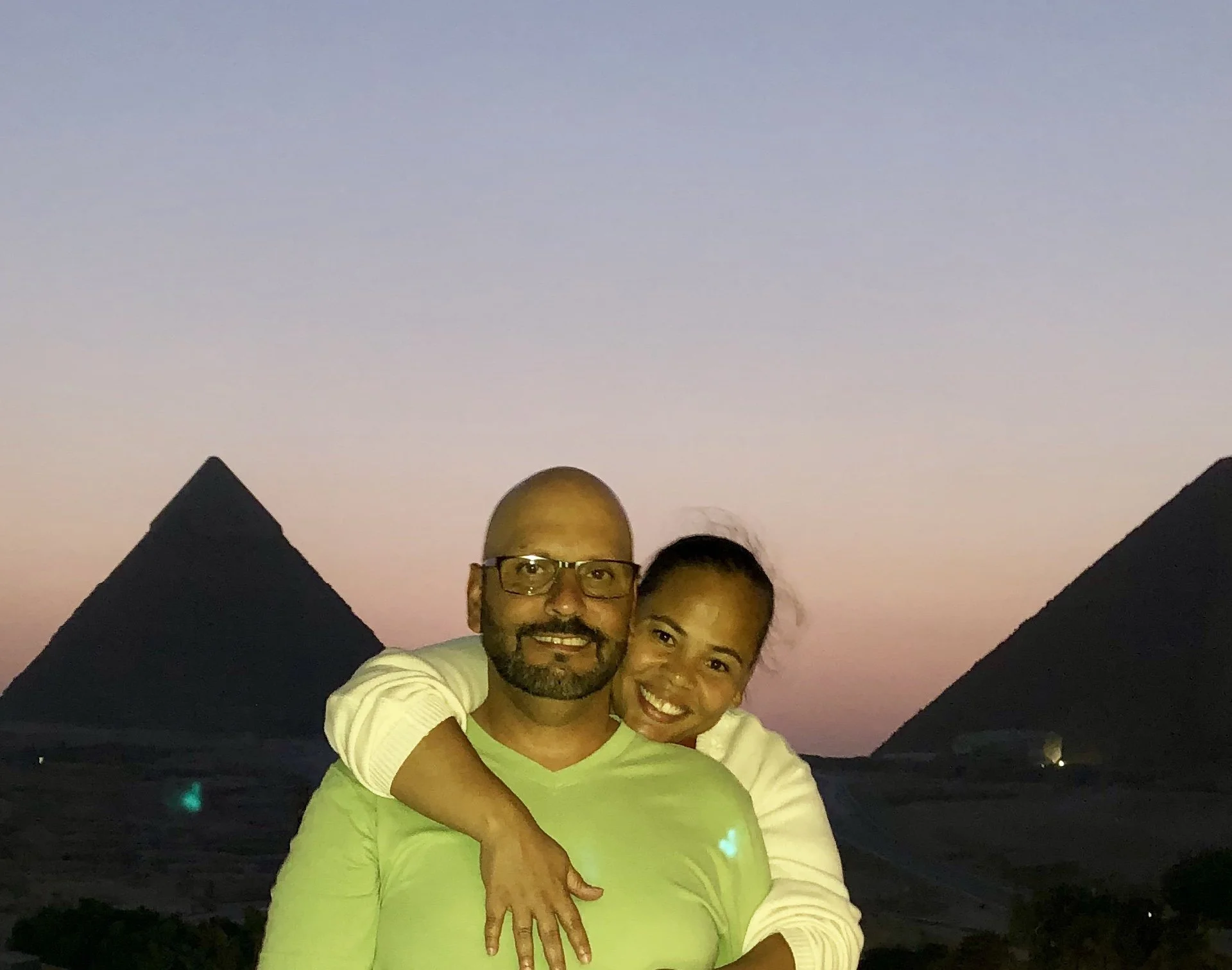 A couple smiling and hugging outdoors during sunset, with pyramids in the background and a colorful sky.