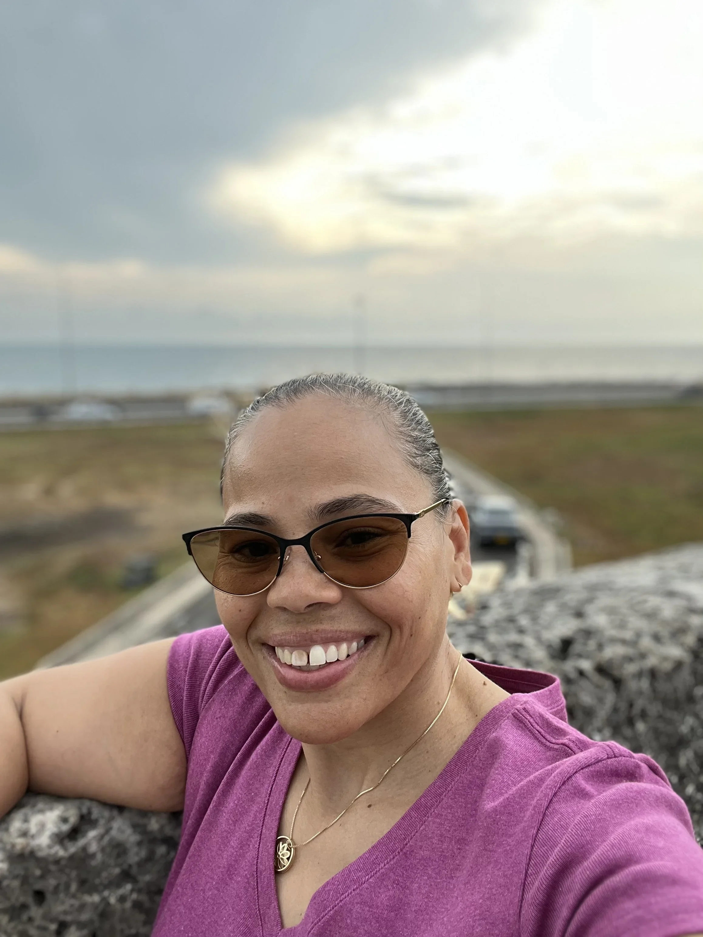 Close-up of a woman smiling with sunglasses, wearing a purple top and a gold pendant necklace, outdoors on a cloudy day with a cloudy sky and landscape in the background.
